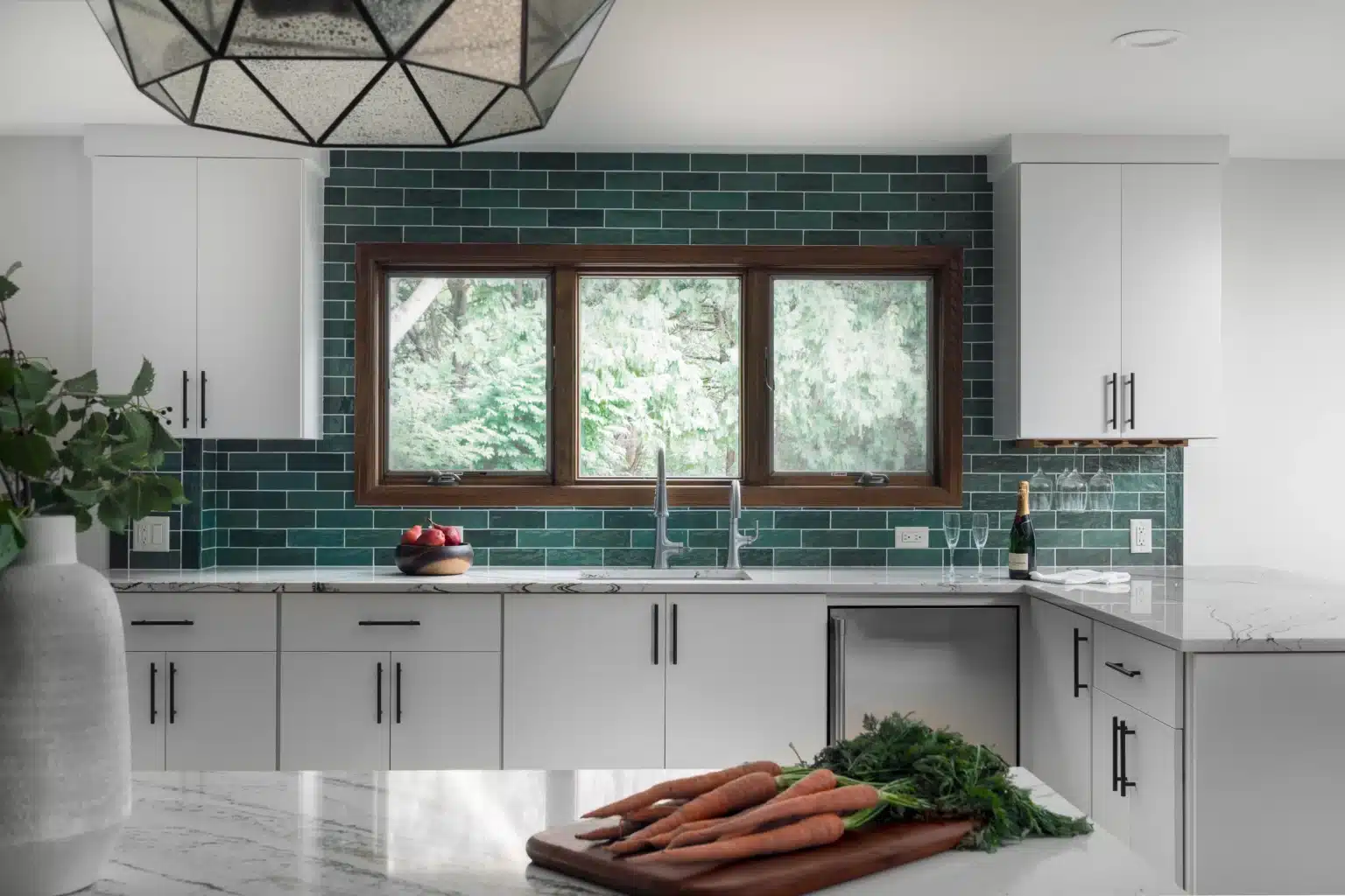 Modern kitchen with green subway tiles, white cabinets, and large window. Carrots on marble countertop, decorative pendant light above, wine bottle nearby.