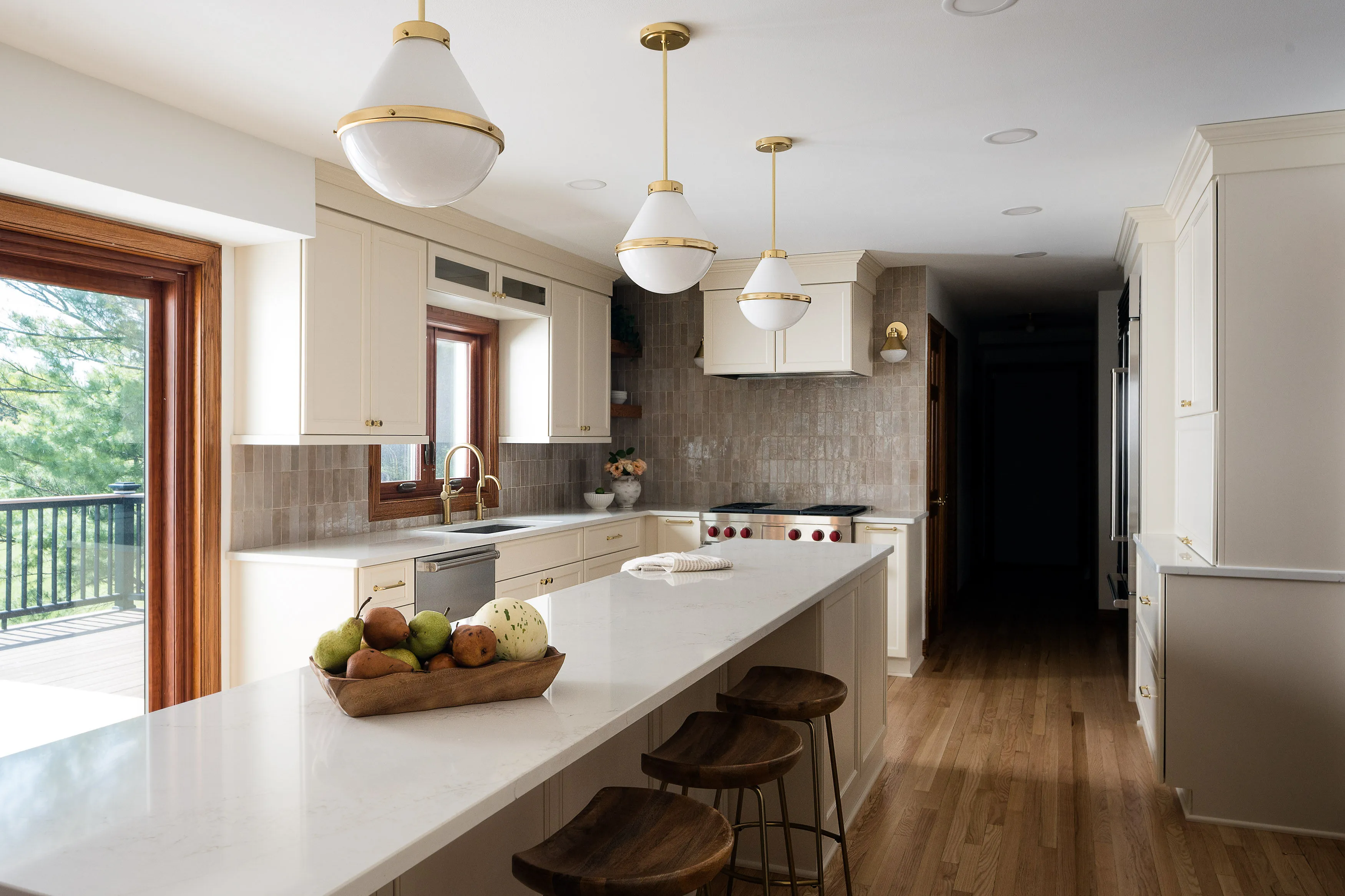 Modern kitchen featuring a long marble island, wooden stools, pendant lighting, and large windows. A fruit bowl decorates the countertop.
