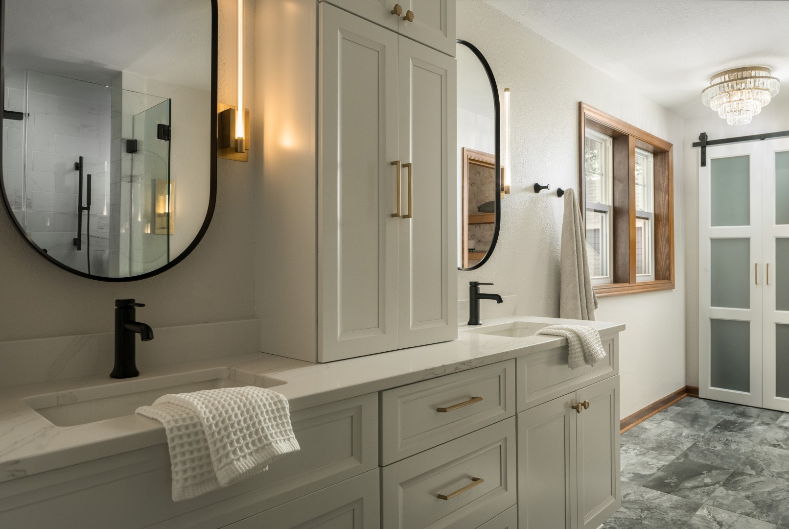 Elegant bathroom with double vanity, black fixtures, oval mirrors, marble countertop, chandelier, frosted sliding door, and large windows. Light gray cabinetry adds elegance.