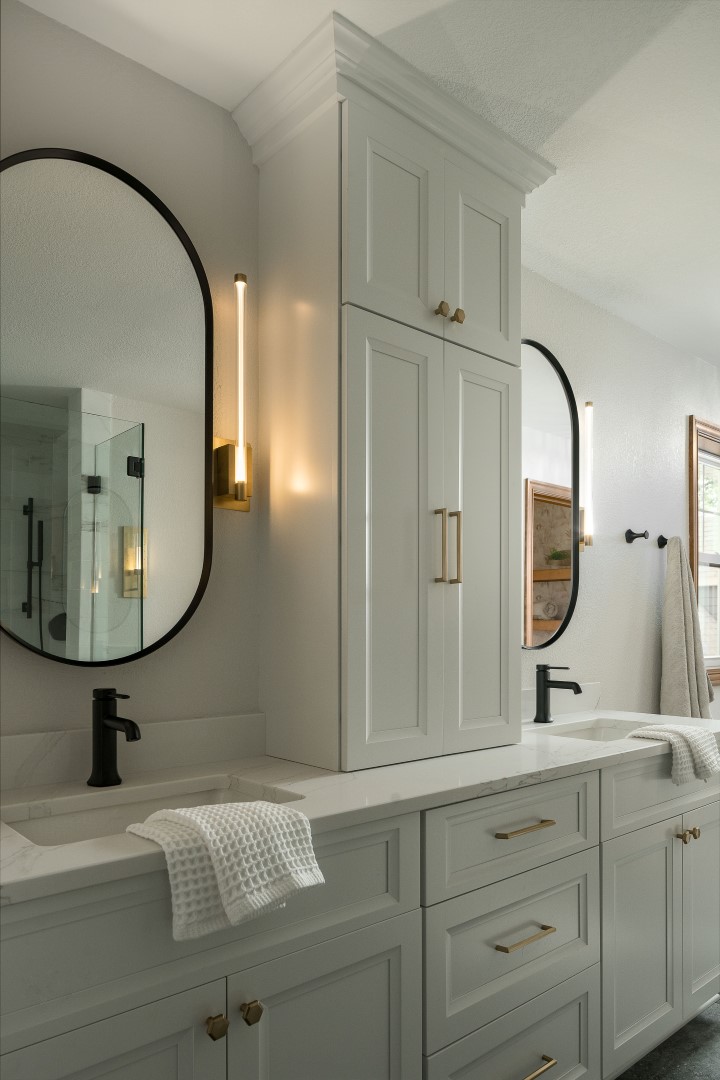 Modern bathroom with dual sinks, black fixtures, and oval mirrors. White cabinetry and warm lighting create a clean, contemporary atmosphere.