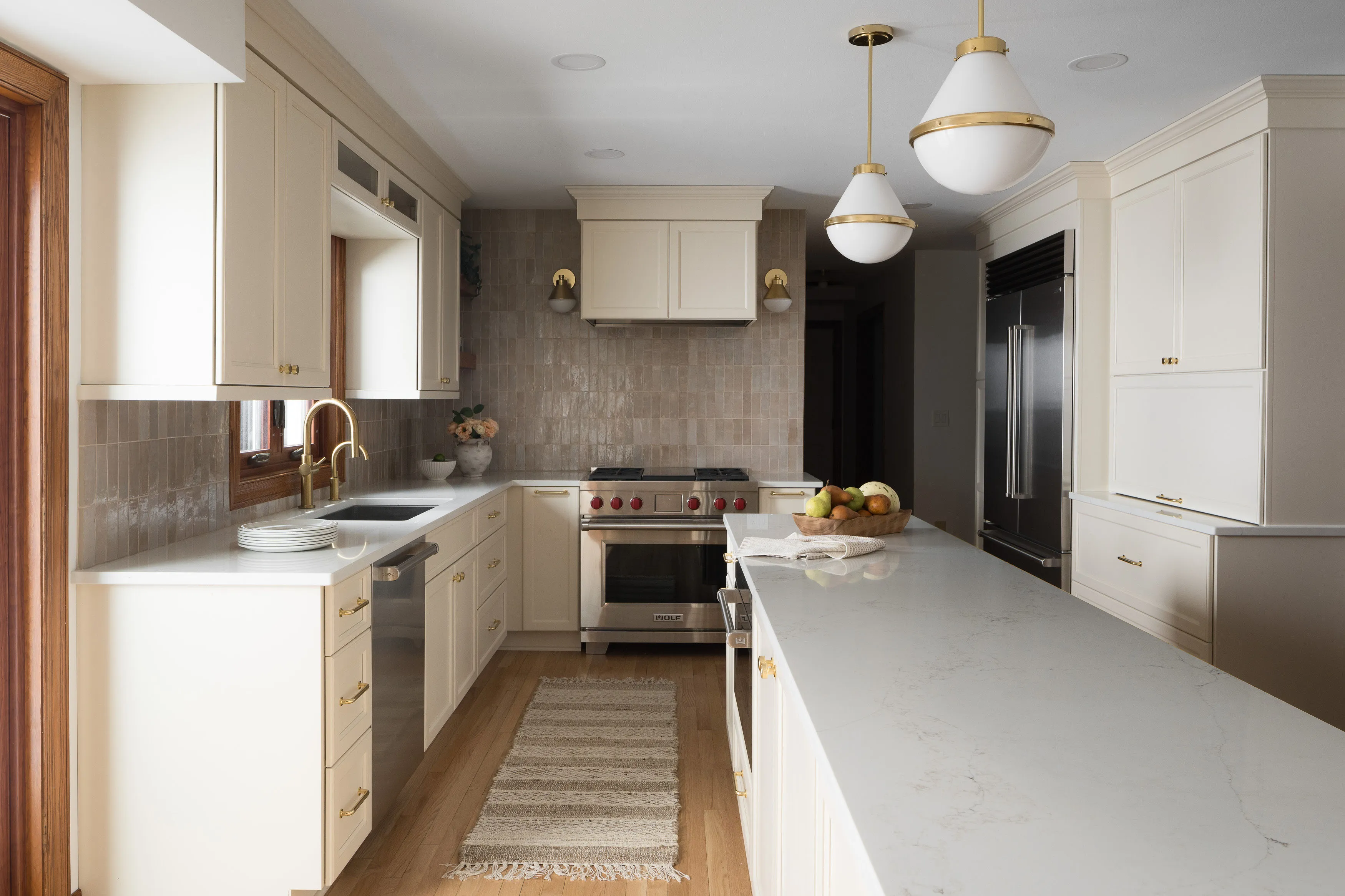 A modern kitchen with beige cabinets, marble countertops, stainless steel appliances, and pendant lighting. A bowl of fruit sits on the island.