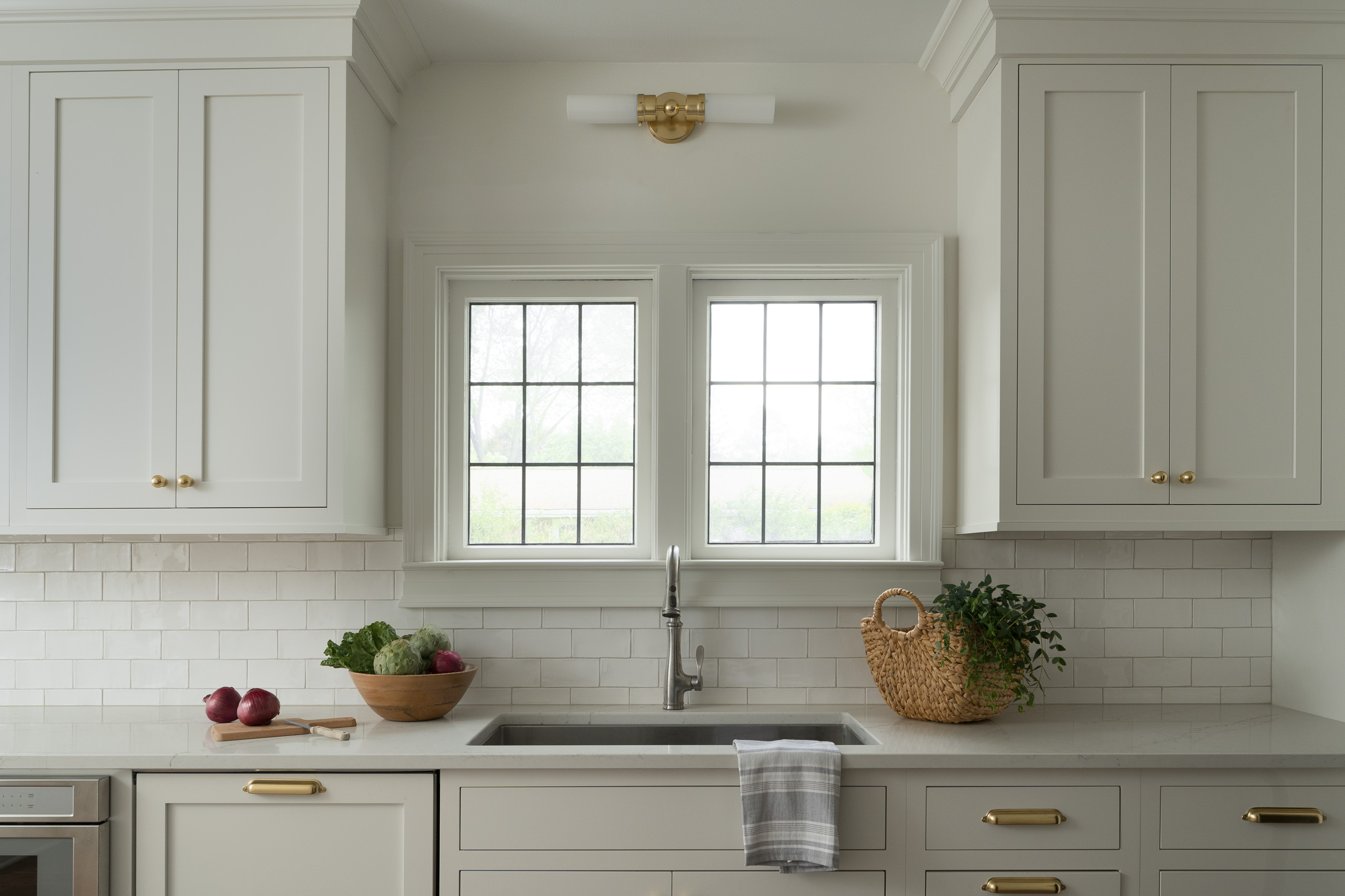 A bright kitchen with white cabinets, gold hardware, two windows, a sink, vegetables in a bowl, and a potted plant.