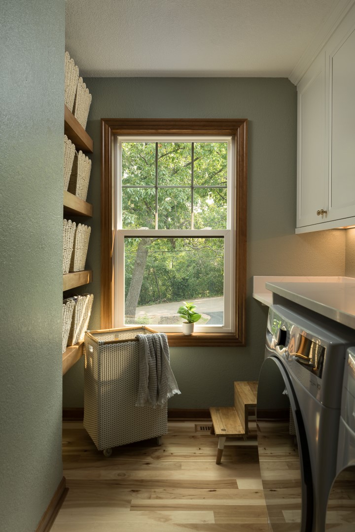 A cozy laundry room with a washing machine, wicker baskets, and a window overlooking lush greenery. A small potted plant adds charm.