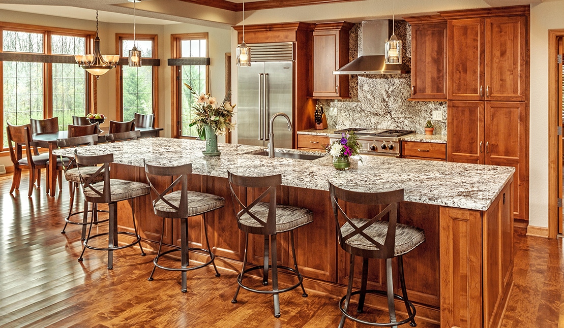 Spacious modern kitchen with wooden cabinets, granite countertops, and barstools. Large windows provide natural light. Dining table with chairs in the background.