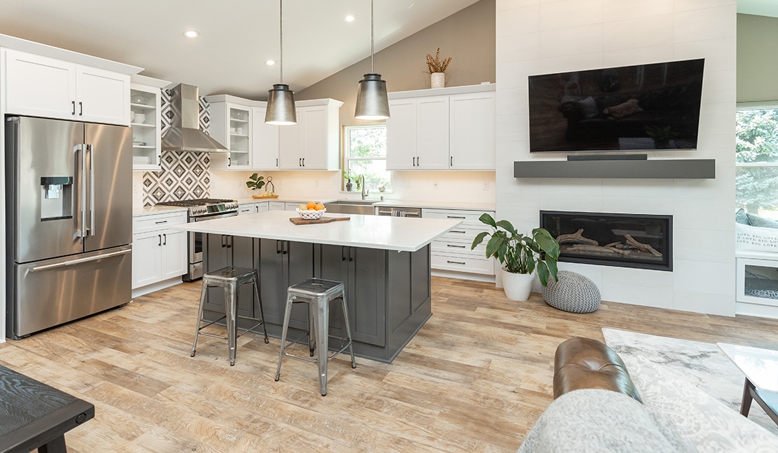 Modern kitchen with white cabinets, gray island, stainless steel appliances, and wood flooring. Features pendant lights, fireplace, and green plants for decor.