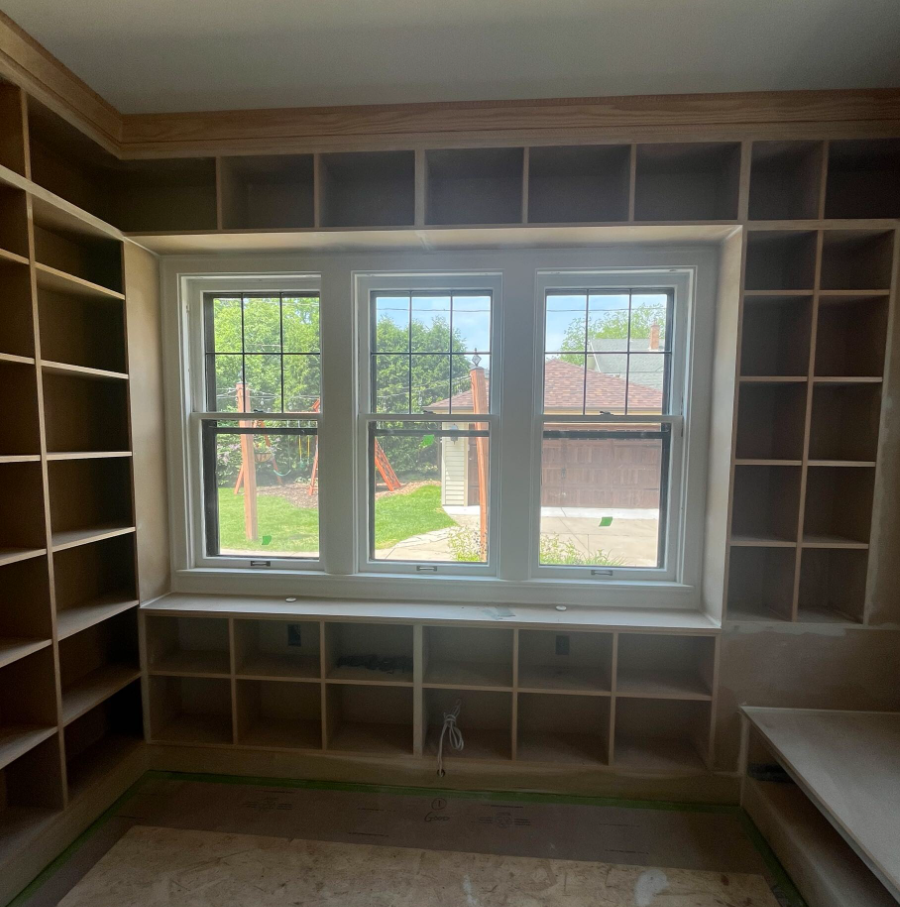 Room with unfinished wooden shelves surrounding a large window, overlooking a backyard with green lawn and a swing set.