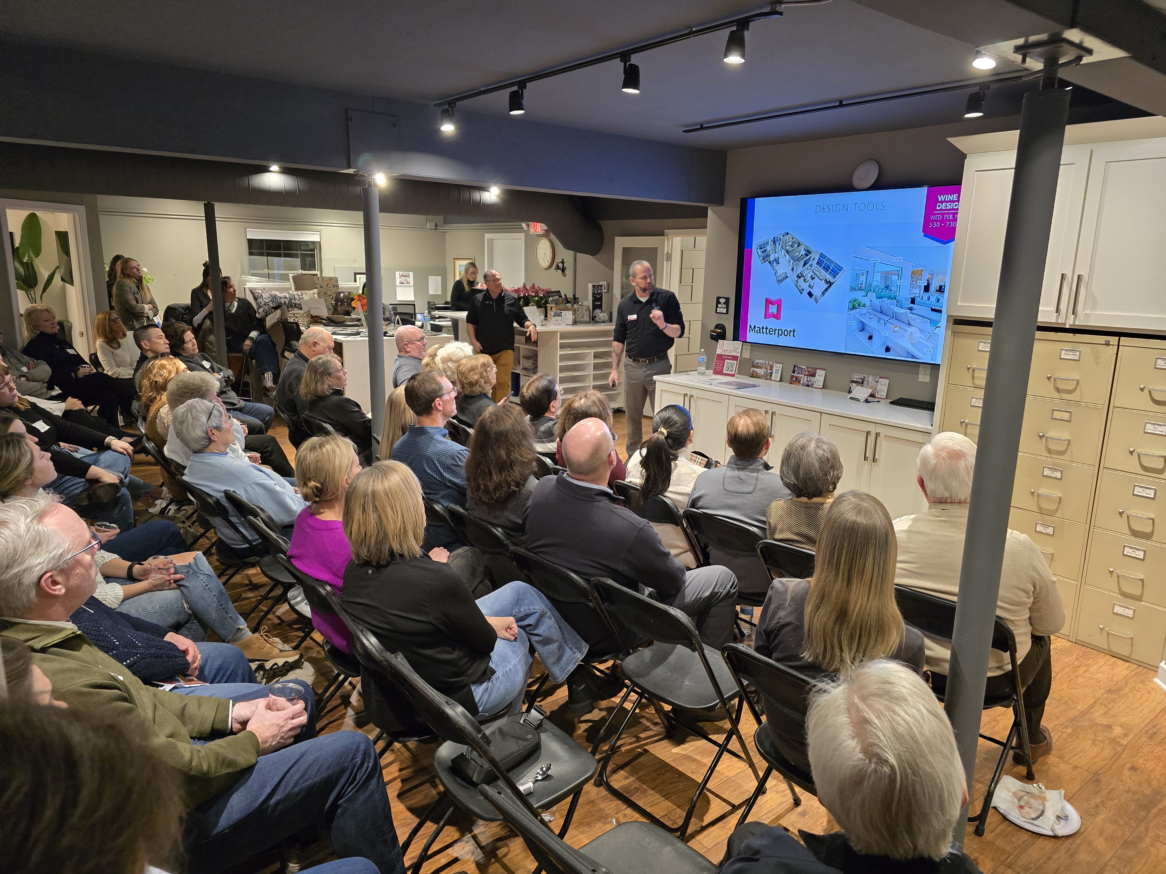 A group of people attending a presentation in a modern room, focused on a screen showing design tools labeled "Matterport."