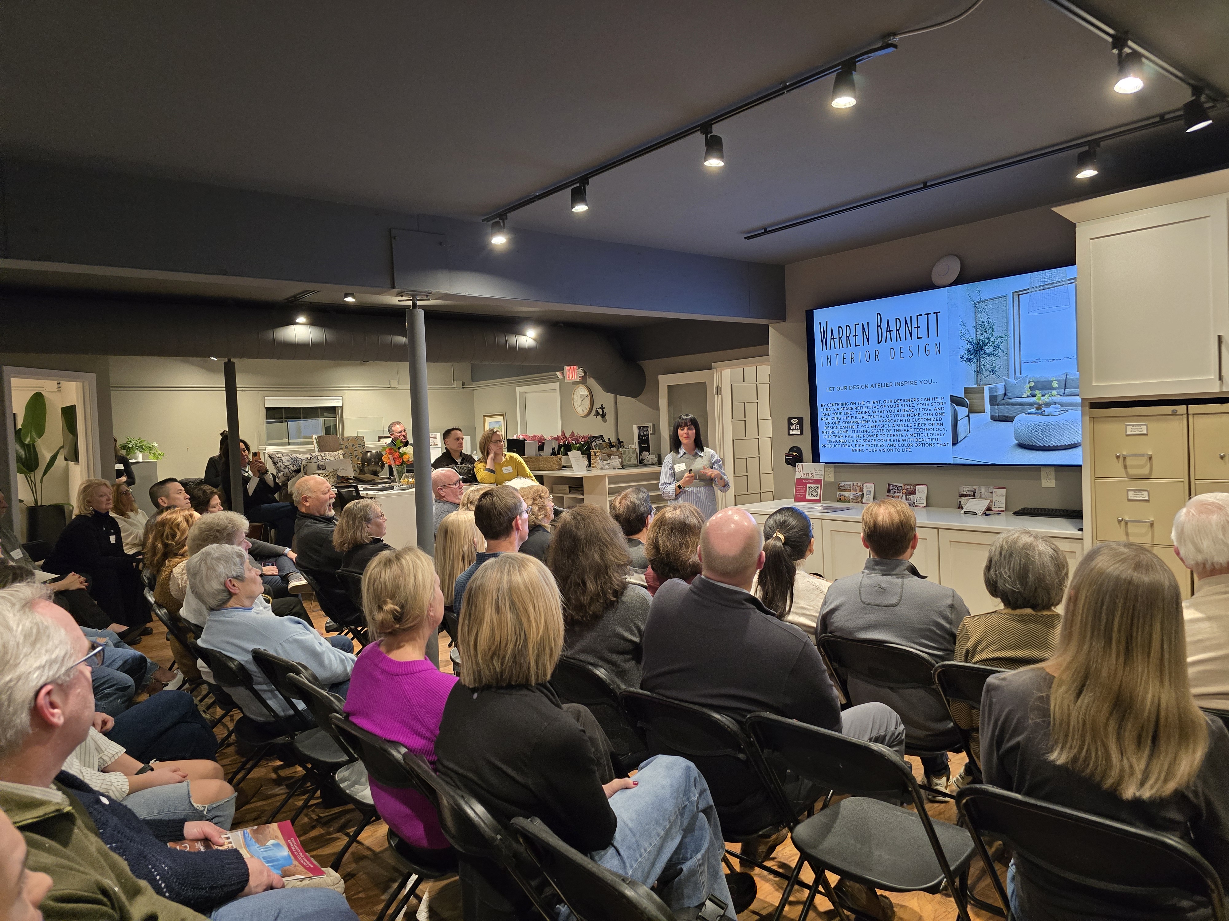 A person presents interior design concepts to an attentive audience in a modern room, featuring a large screen displaying "Warren Barnett Interior Design."