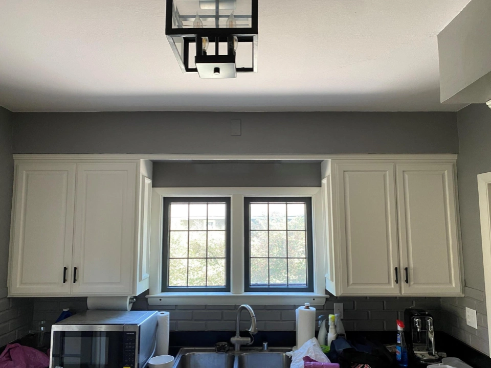 A kitchen with white cabinets, a modern light fixture, and a window above a sink. It includes a microwave and cleaning supplies.