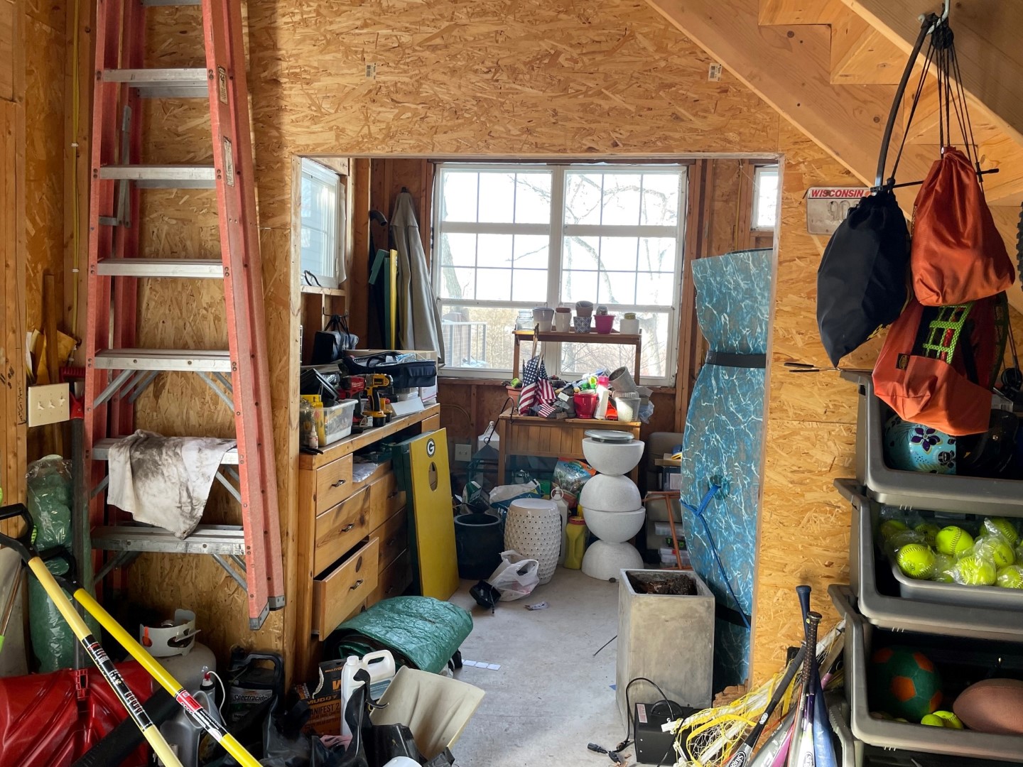 A cluttered garage with tools, sports equipment, American flags, and storage bins. A red ladder and window with daylight are visible.