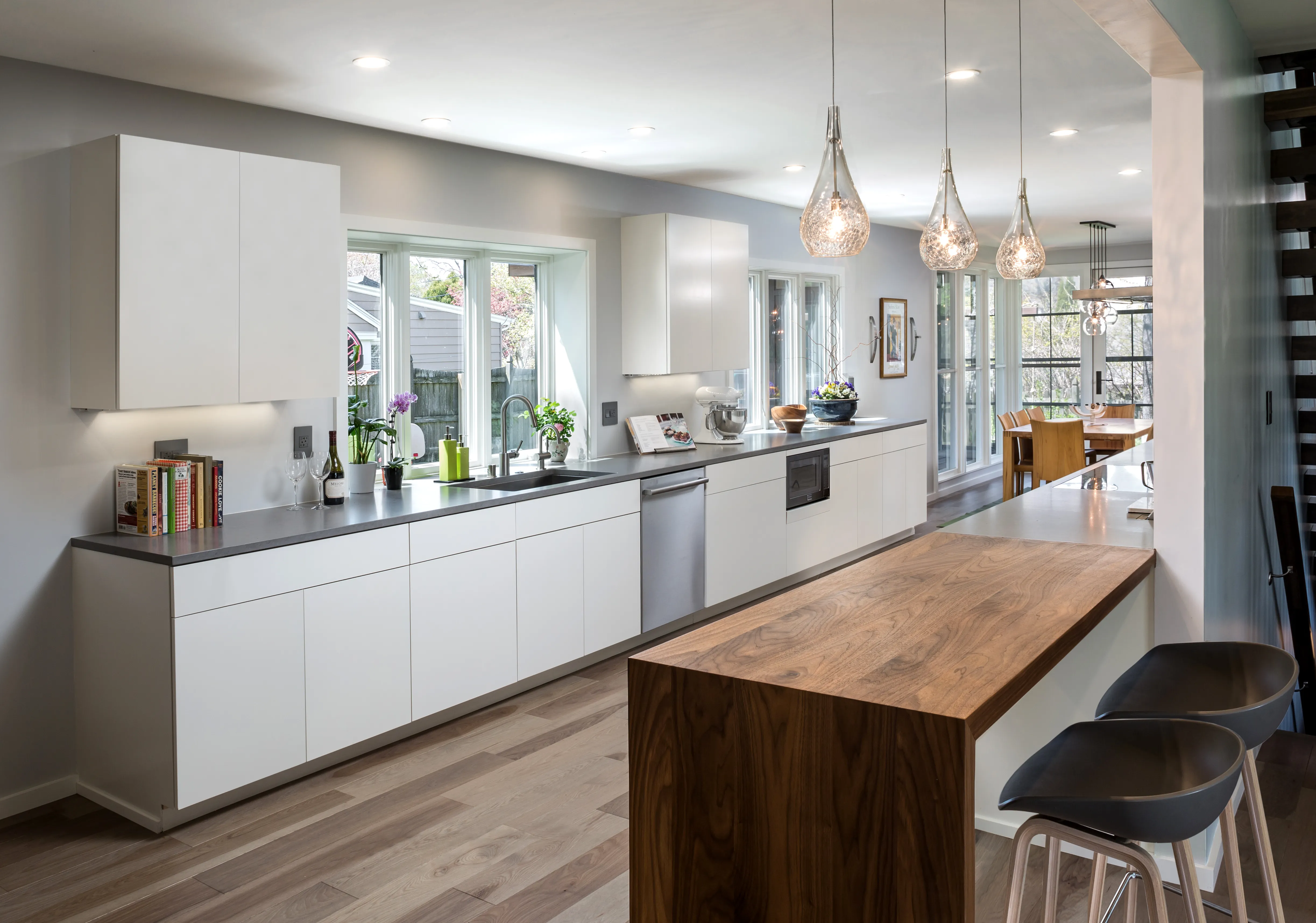 Modern kitchen with white cabinets, wooden island, and pendant lights. Bright, airy space featuring large windows, plants, and a dining area in the background.