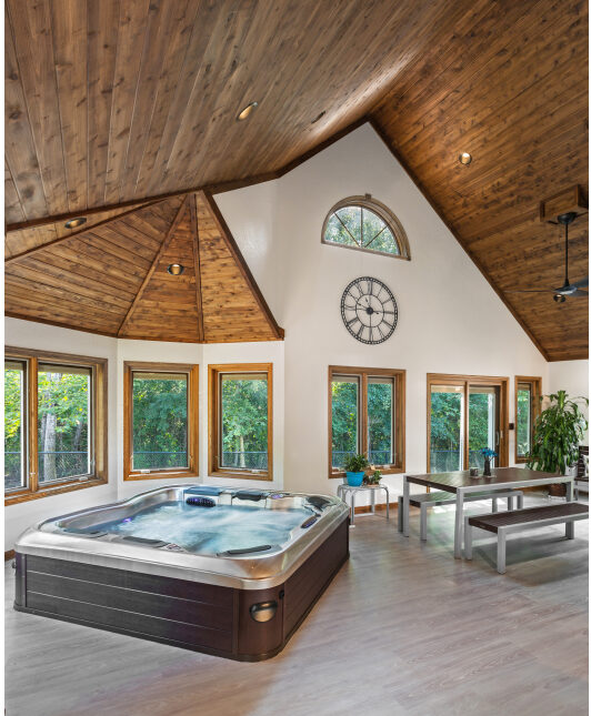 A new in-home spa built by S.J. Janis. Vauleted wood-grain ceilings, light wood floors, wall of wood-framed windows in white wall, hot tub in front of the windows.