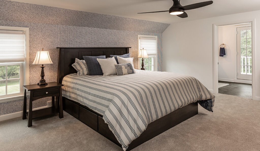 Bedroom in whole-house remodel. Black bed frame with drawers underneath, beige carpet, two double-hung white windows, gray wall, black ceiling fan.