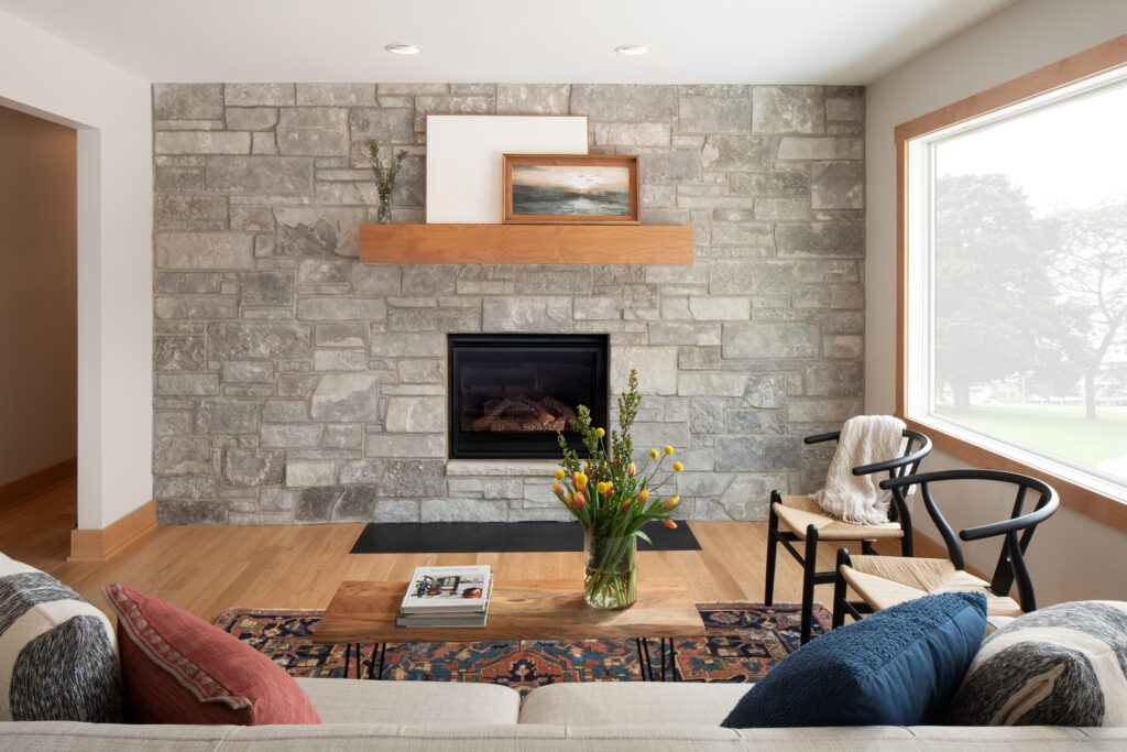 Renovated living room featuring a new, in-wall fireplace in a stone wall. and new light wood flooring. Large picture window on right.