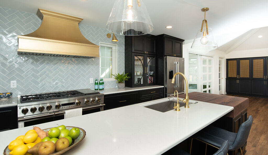 Remodeled kitchen featuring a new kitchen island with white stone counter, matte gold hood range, stainless steel refrigerator, and new lighting fixtures. Pale blue-gray herringbone tile backsplash.