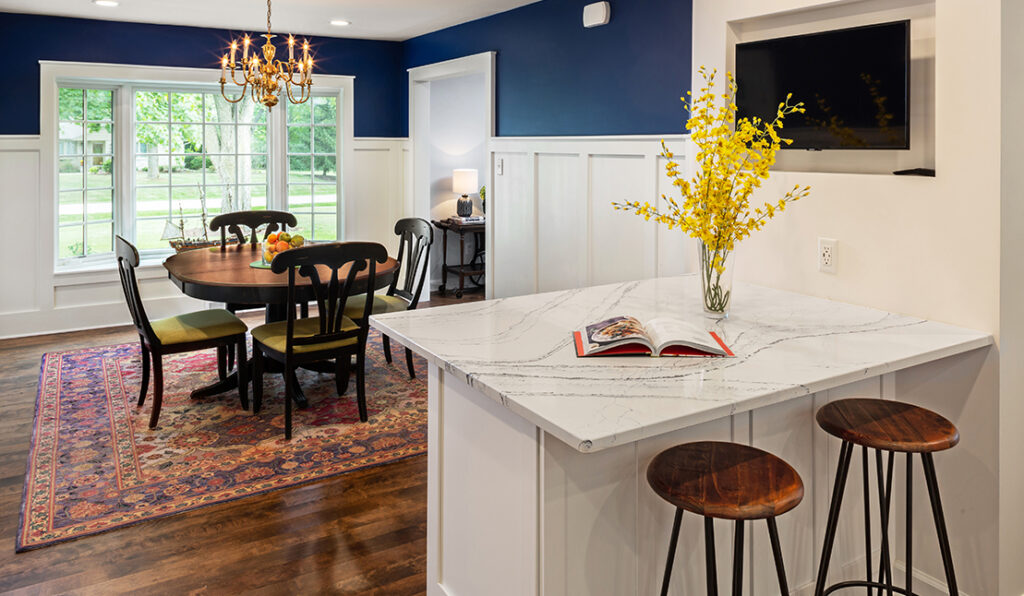 Newly renovated kitchen and dining room with new white cabinetry, small peninsula with white marble countertop and two stools, wood flooring, open to dining area with navy blue walls and white wainscoting, and large picture window with grids.