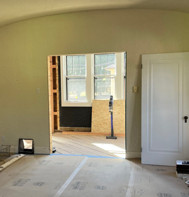 Room under renovation with newly cut doorway, exposed wooden frame, and windows. A vacuum stands in the adjacent room on wooden floorboards.