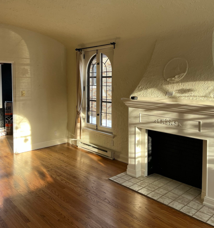 Sunlit room with arched window, graceful curtains, and an elegant fireplace against textured walls. Warm wooden floor enhances the cozy, serene ambiance.