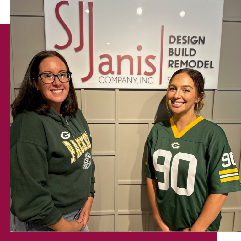 Two people in Green Bay Packers attire stand smiling in front of a sign for S.J. Janis Company, Inc., a design firm.