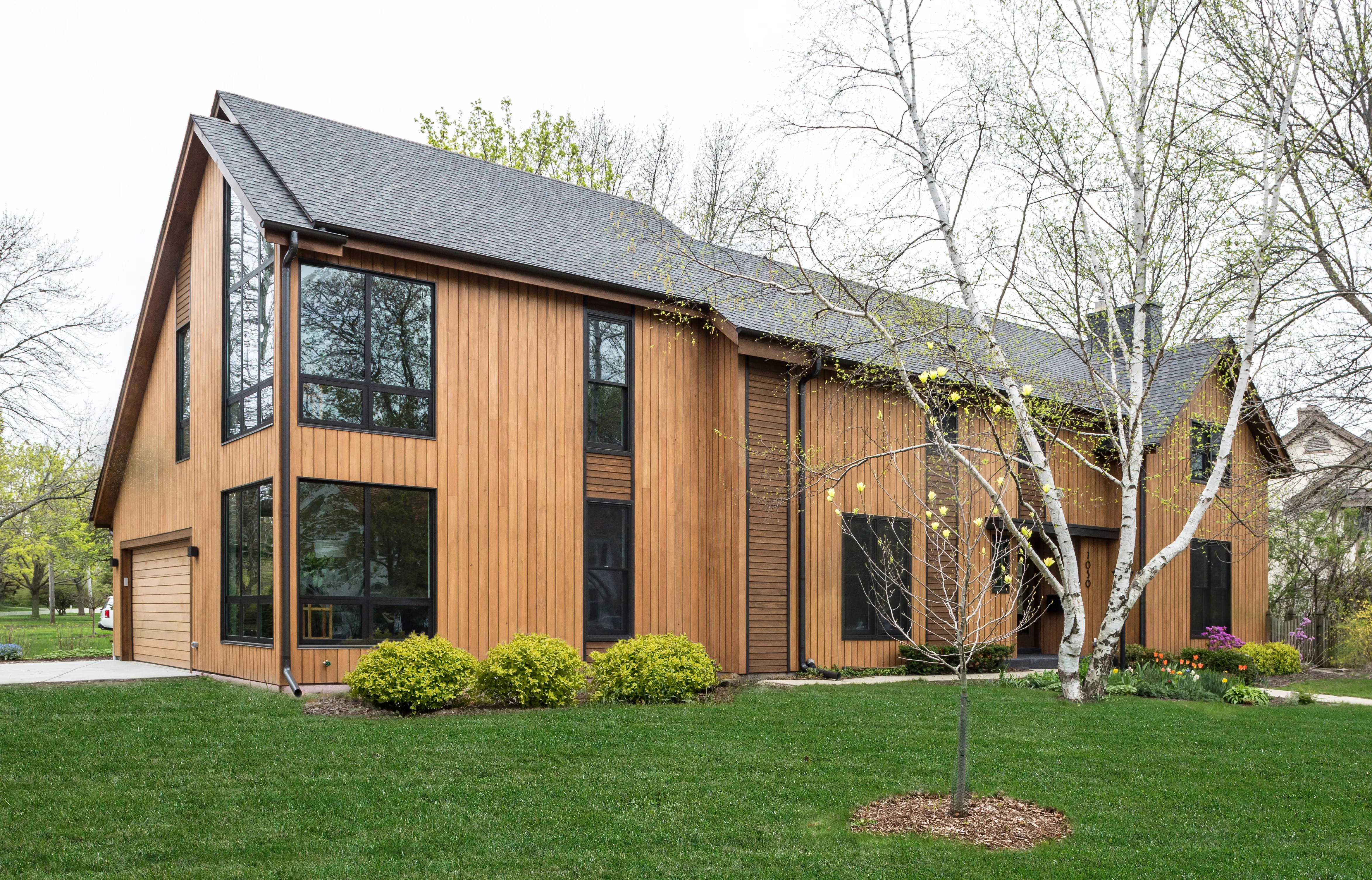 Modern wooden house with large windows, surrounded by green lawn and trees. Spring blooms and neatly trimmed bushes enhance the landscape.