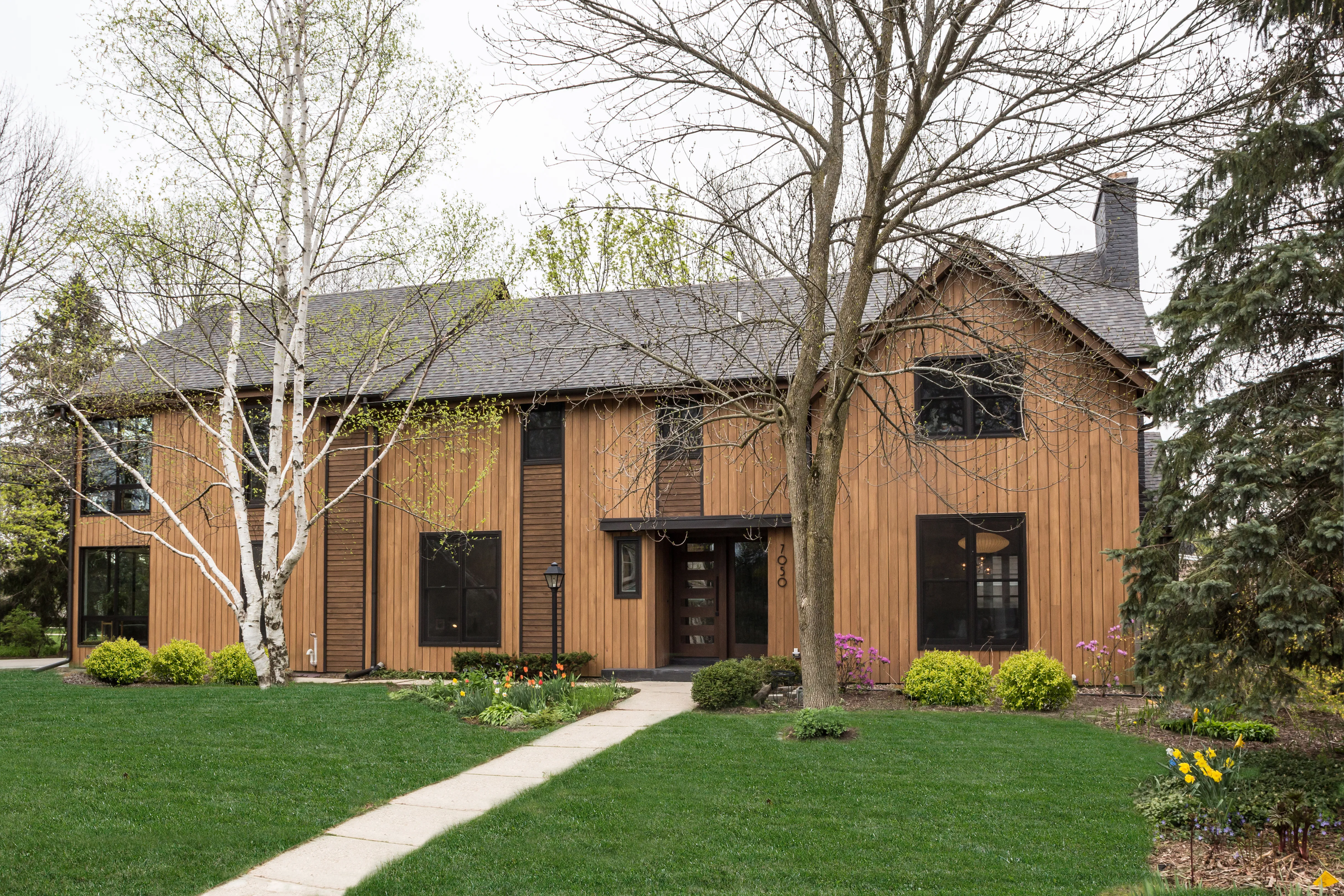 A wooden house with large windows, surrounded by trees and a well-kept garden, showcasing a serene suburban setting.