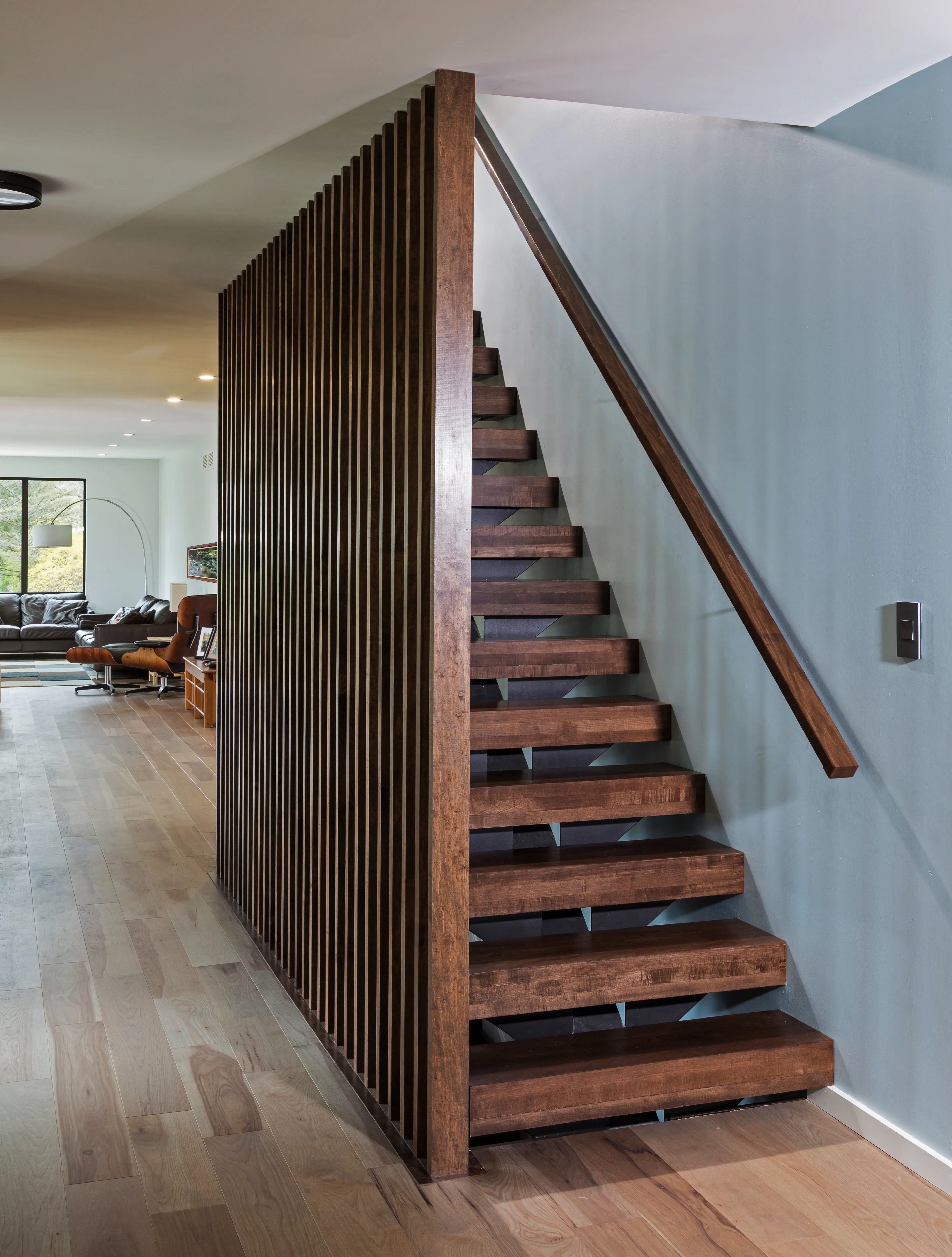 Modern interior with wooden floating staircase, slatted partition, and airy living room. Large windows let in natural light, highlighting minimalist design.