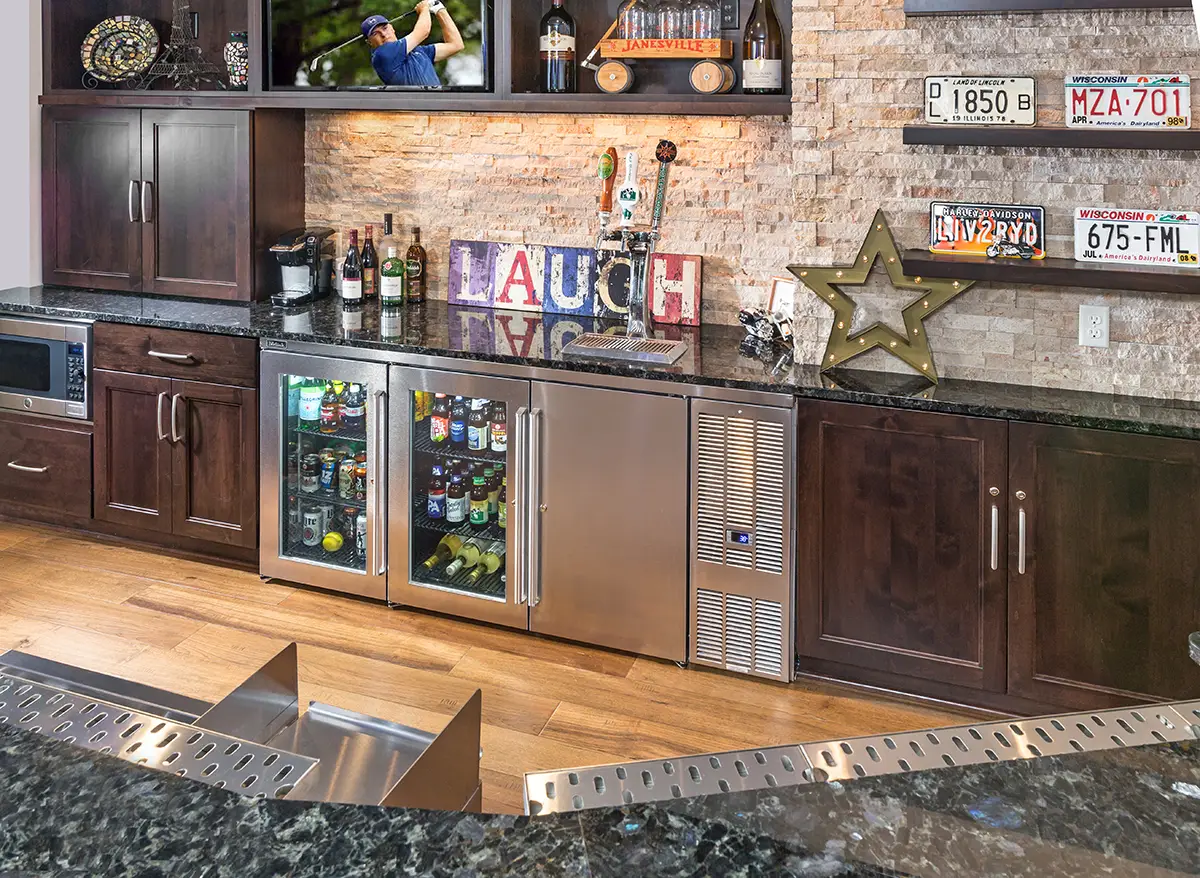 Modern home bar with dark granite countertop, wooden cabinets, and blue pendant lights. Decor includes license plates, a vase, and shelves with bottles.