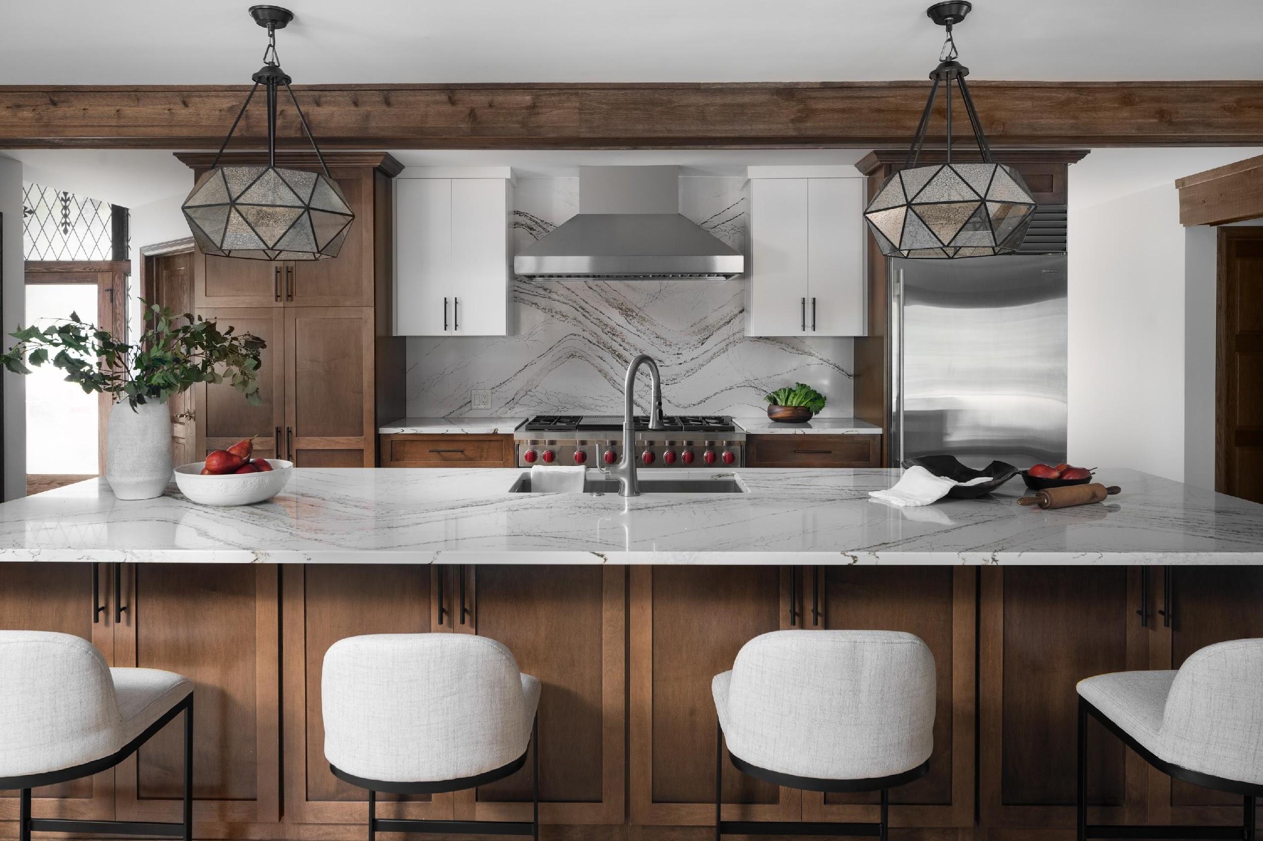 Modern kitchen with marble countertop, wooden cabinets, geometric light fixtures, stainless steel appliances, and elegant decor, featuring white cushioned bar stools.