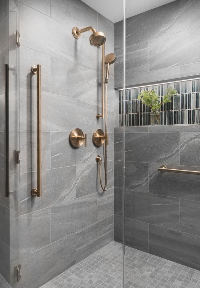 Modern shower with gray tiles, brass fixtures, glass door, and a small shelf holding plants. Sleek, minimalist design in a contemporary bathroom.