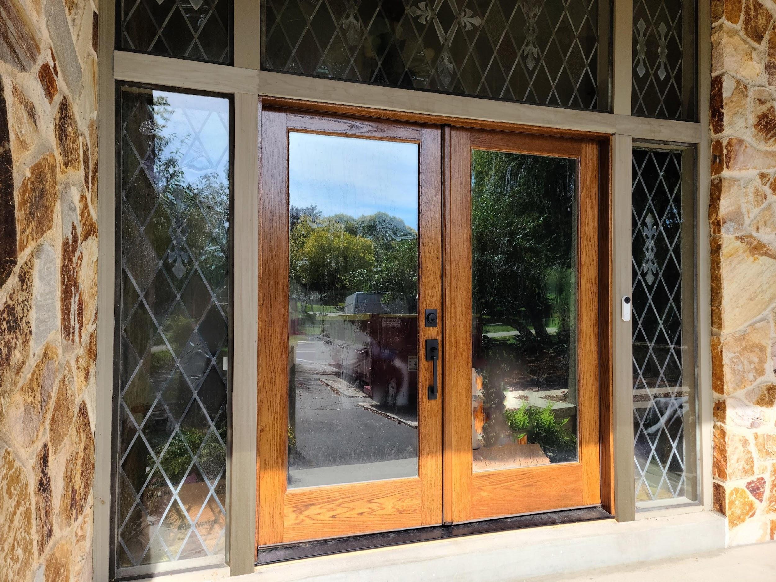 Wooden double door with decorative glass panels and stone wall, reflecting trees and a visible pathway outside.