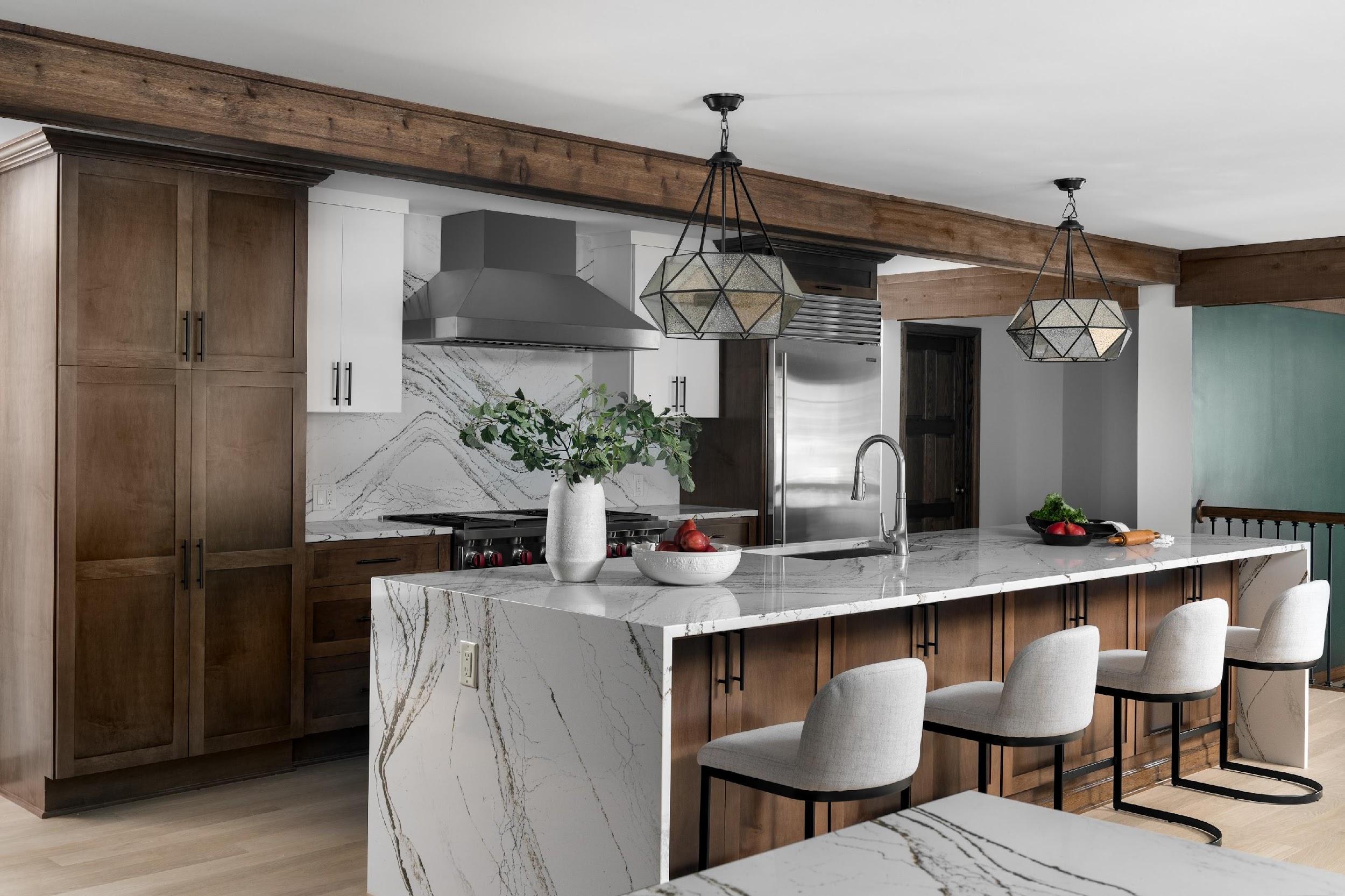 Modern kitchen featuring marble countertops, wooden cabinets, geometric pendant lights, and stainless steel appliances. Four upholstered chairs line the island. No people present.