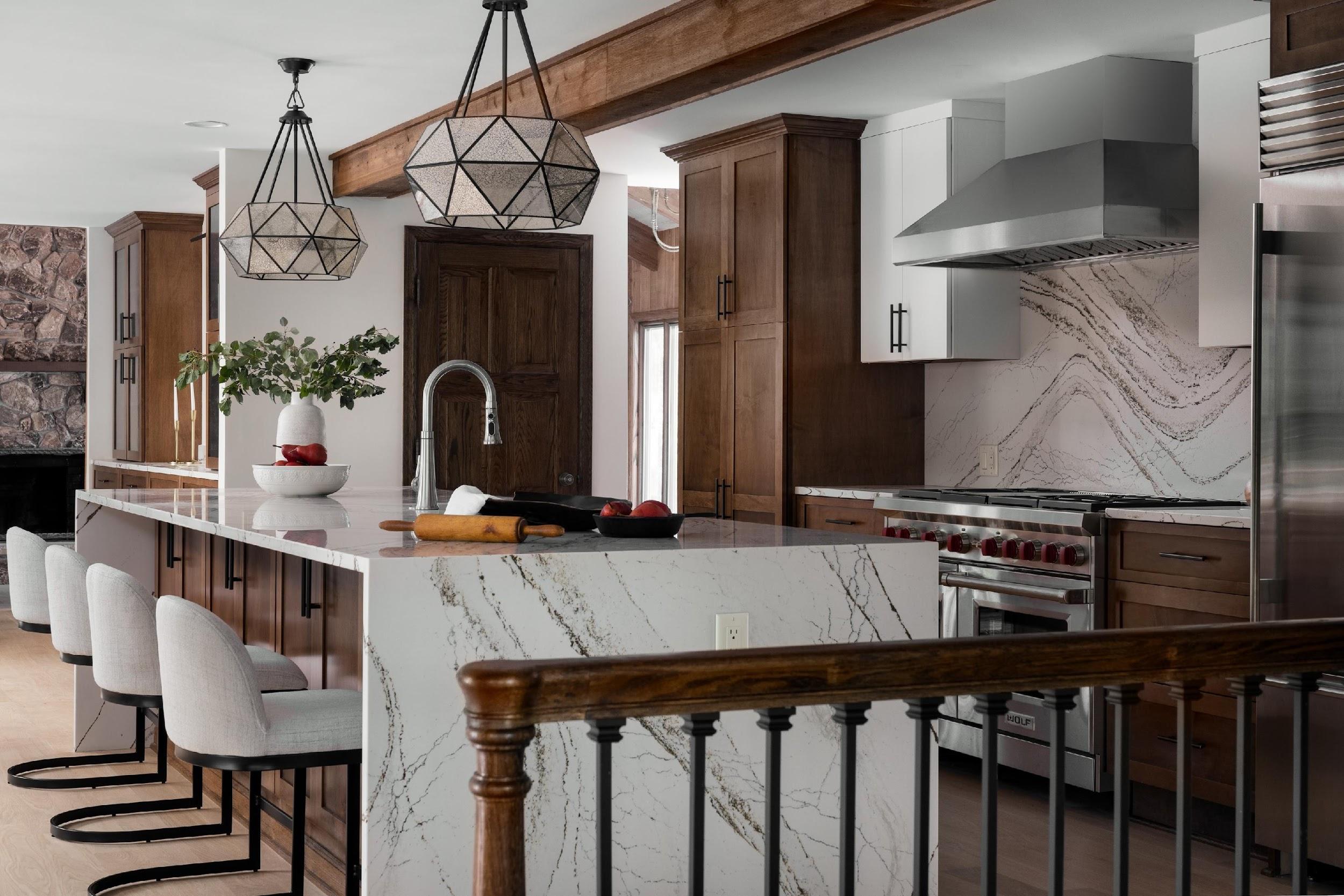Modern kitchen featuring marble countertops, wooden cabinets, geometric light fixtures, and elegant seating. Stainless steel appliances and a decorative plant complete the space.