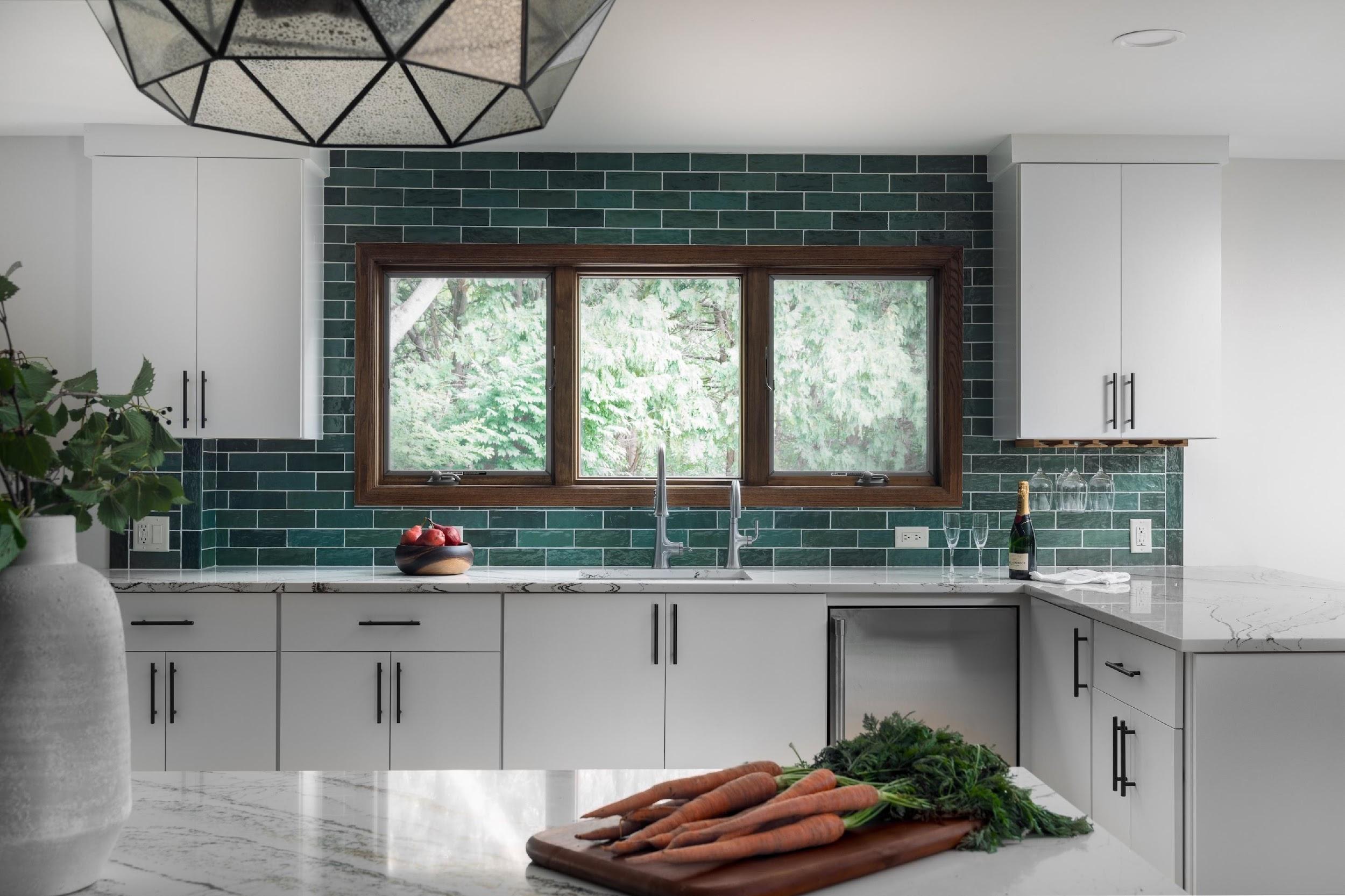 Modern kitchen with green tiled backsplash, white cabinets, and marble countertop. Carrots on cutting board; bowl of apples, champagne, and glasses visible.
