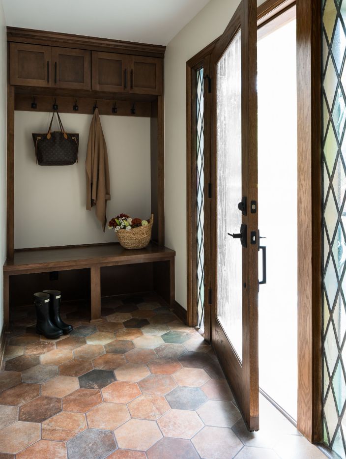 A cozy entryway featuring a bench, coat hooks, a basket, boots, and a wooden door with decorative glass panels, tiled floor.