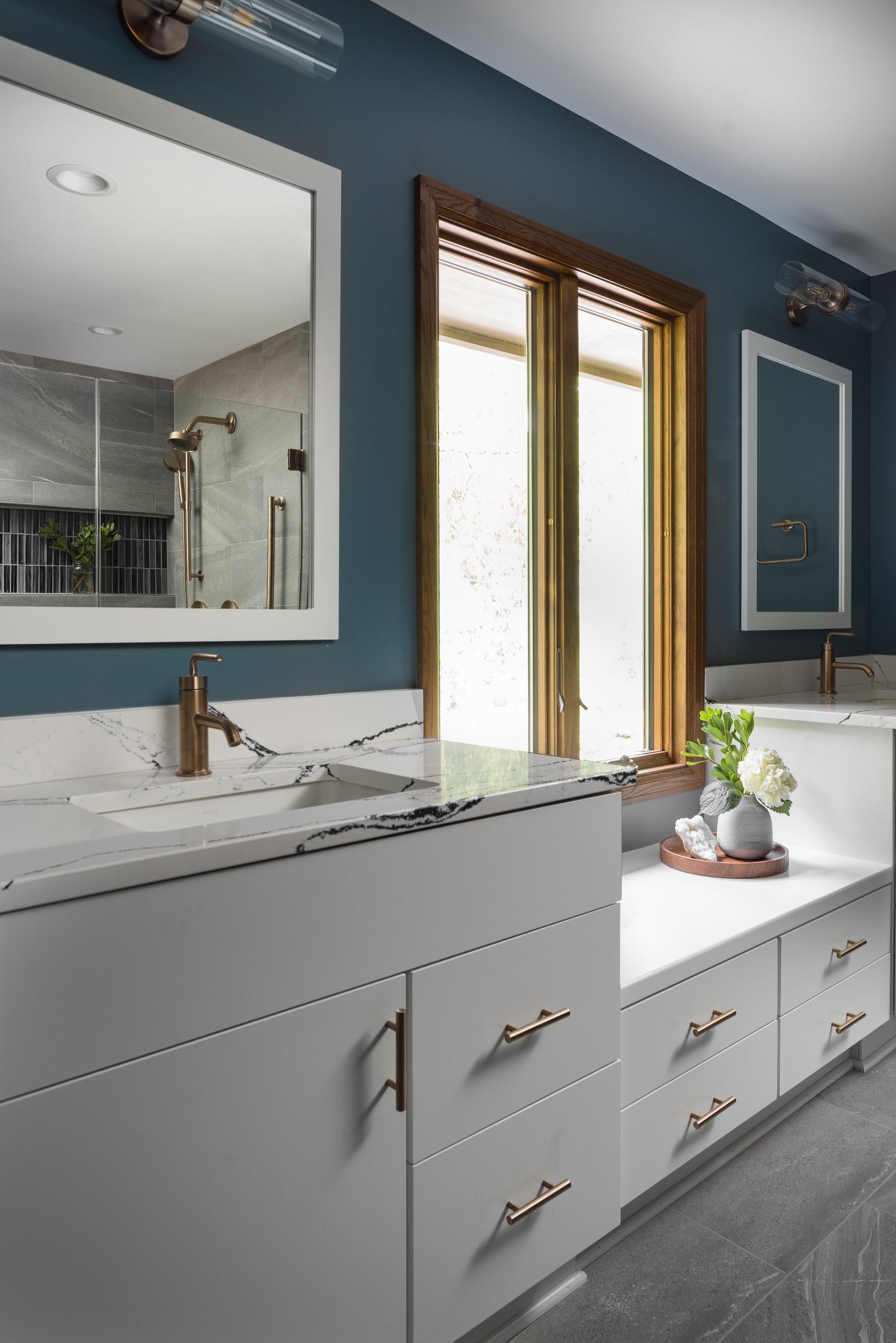 Modern bathroom with dual sinks, marble countertops, brass faucets, teal walls, large mirror, and a small plant by the window. Sleek, elegant design.