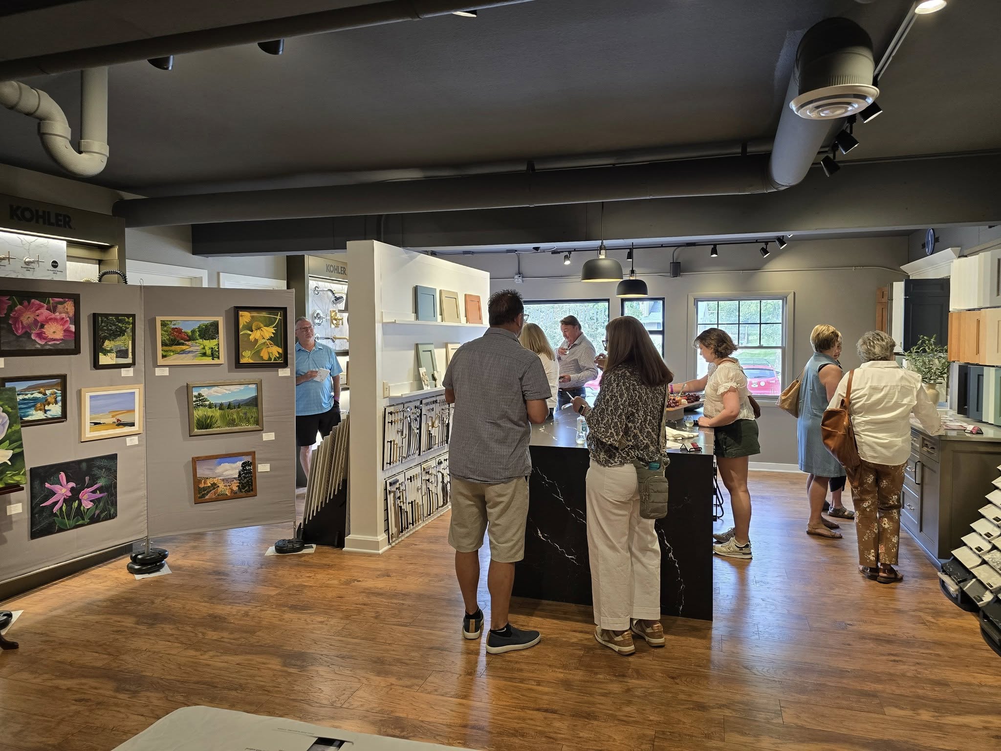 Several people in a gallery viewing paintings and discussing. The space has wooden floors and displays various art pieces on the walls.
