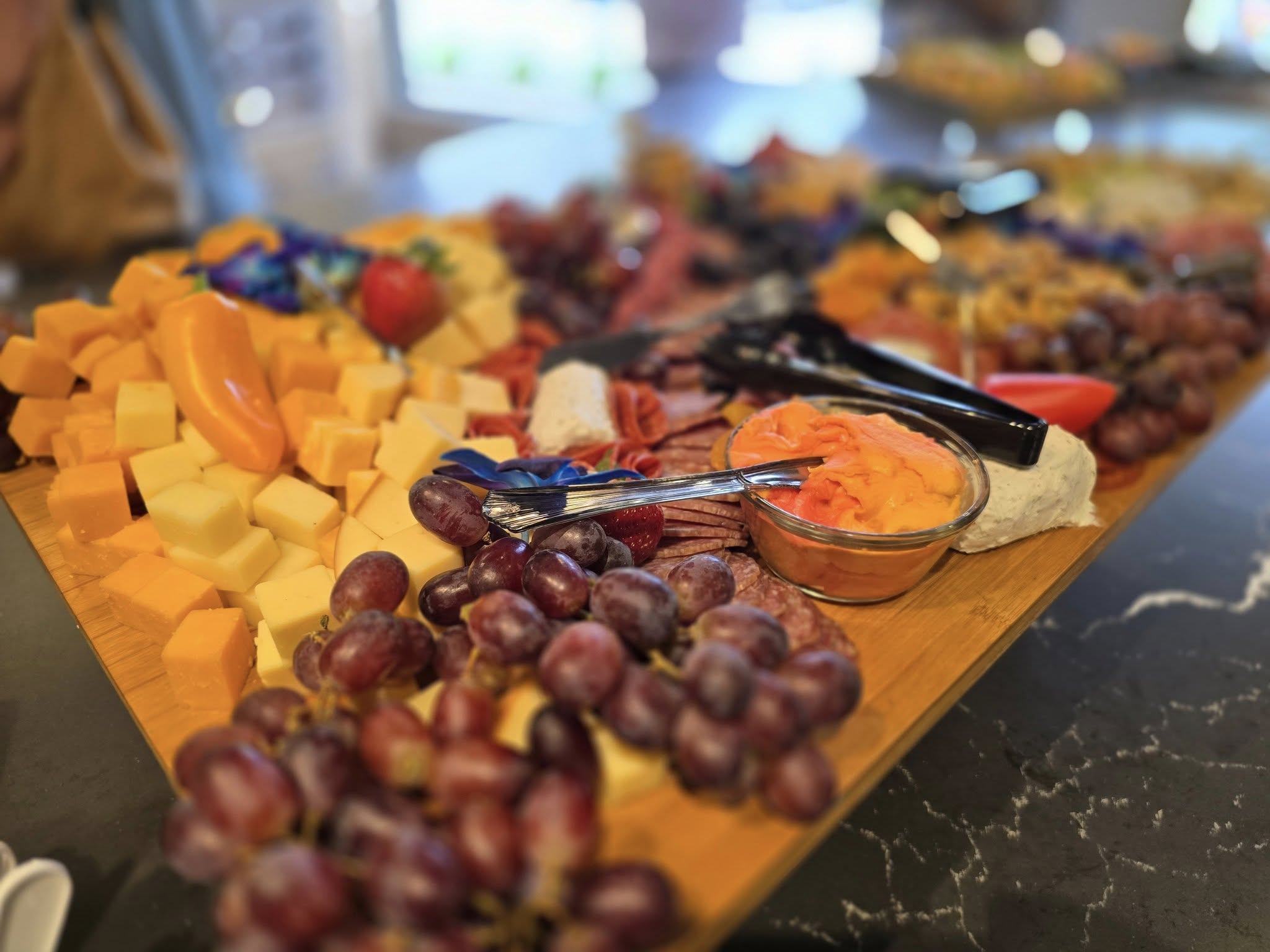 Cheese and fruit platter with grapes, cheddar cubes, sliced meats, and dips on a wooden board in a kitchen setting.