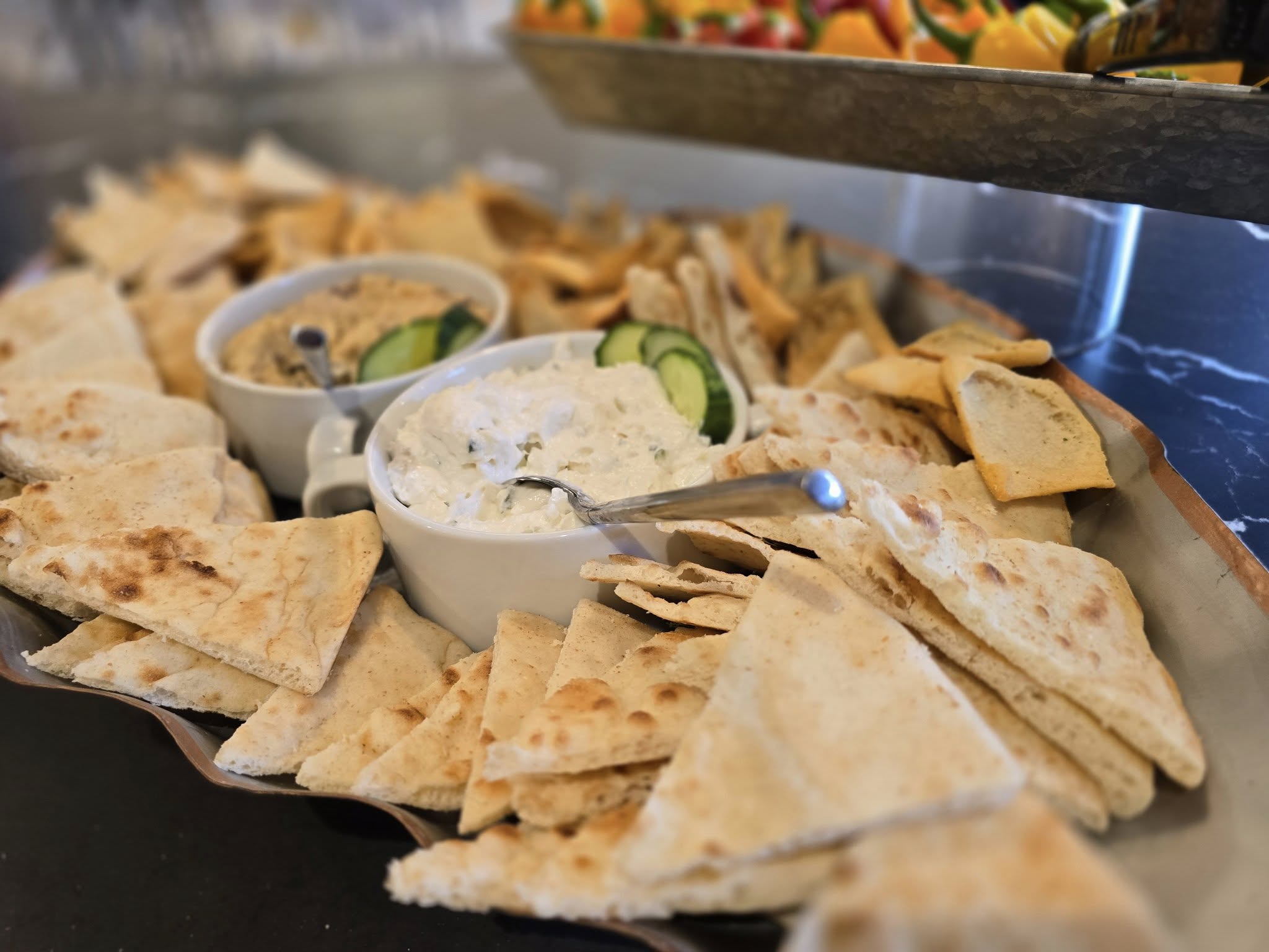 A platter of pita bread with creamy dips, garnished with cucumber slices, arranged elegantly on a table with a colorful fruit tray.