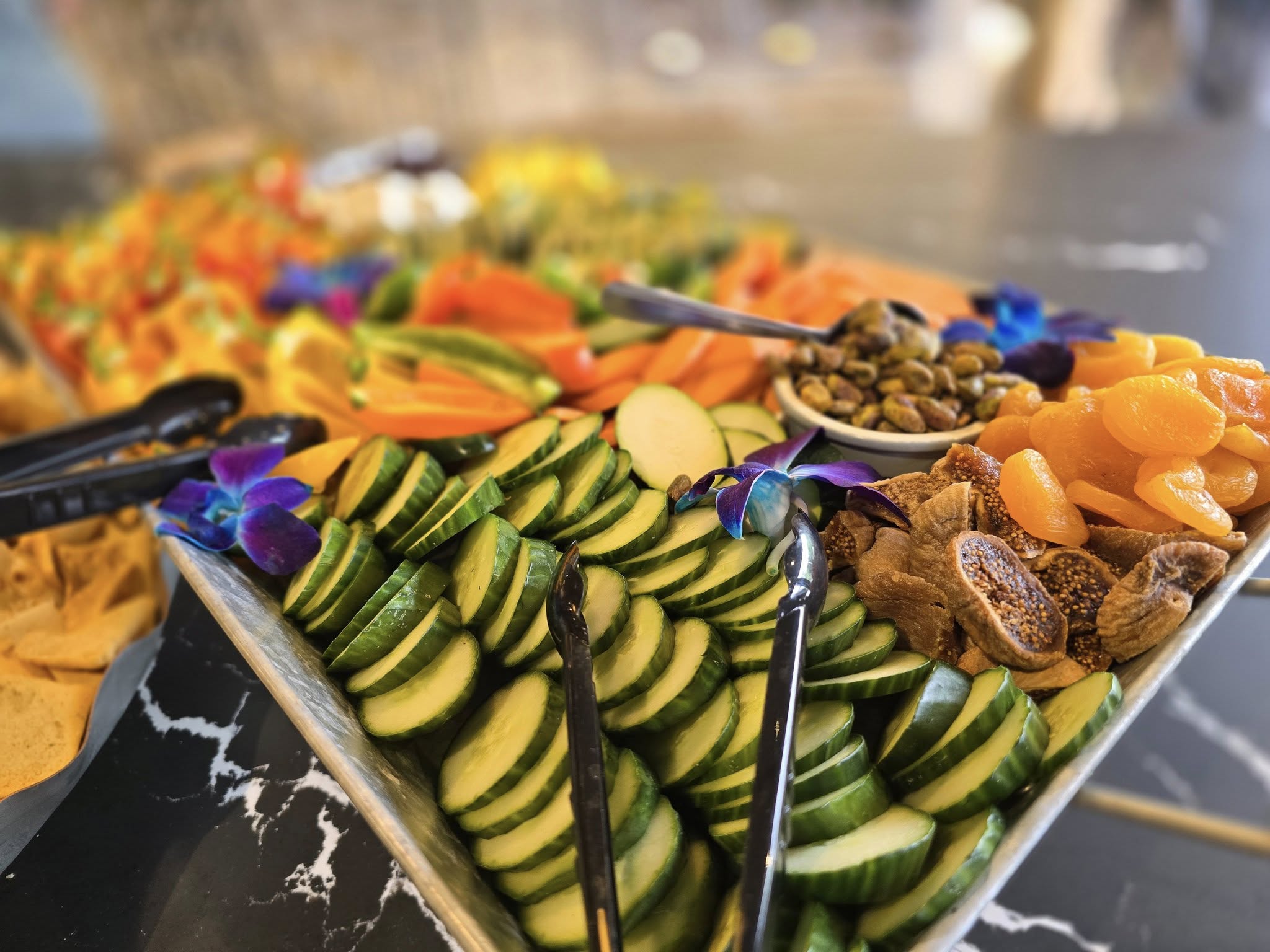 A vibrant platter featuring sliced cucumbers, carrots, pistachios, dried apricots, figs, and blue flowers on a marble table with tongs.
