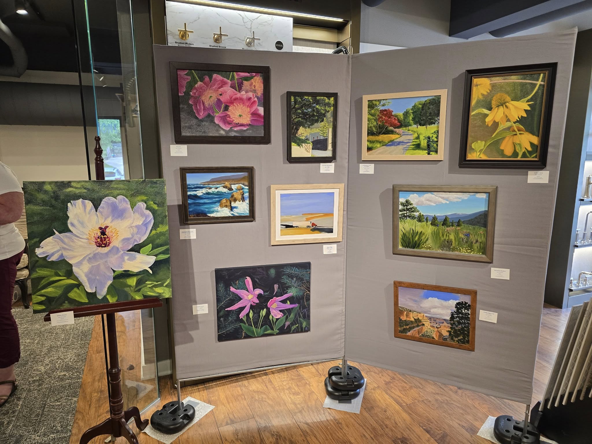 An art display features various colorful landscape and floral paintings on a panel in a gallery setting, with a person partially visible.