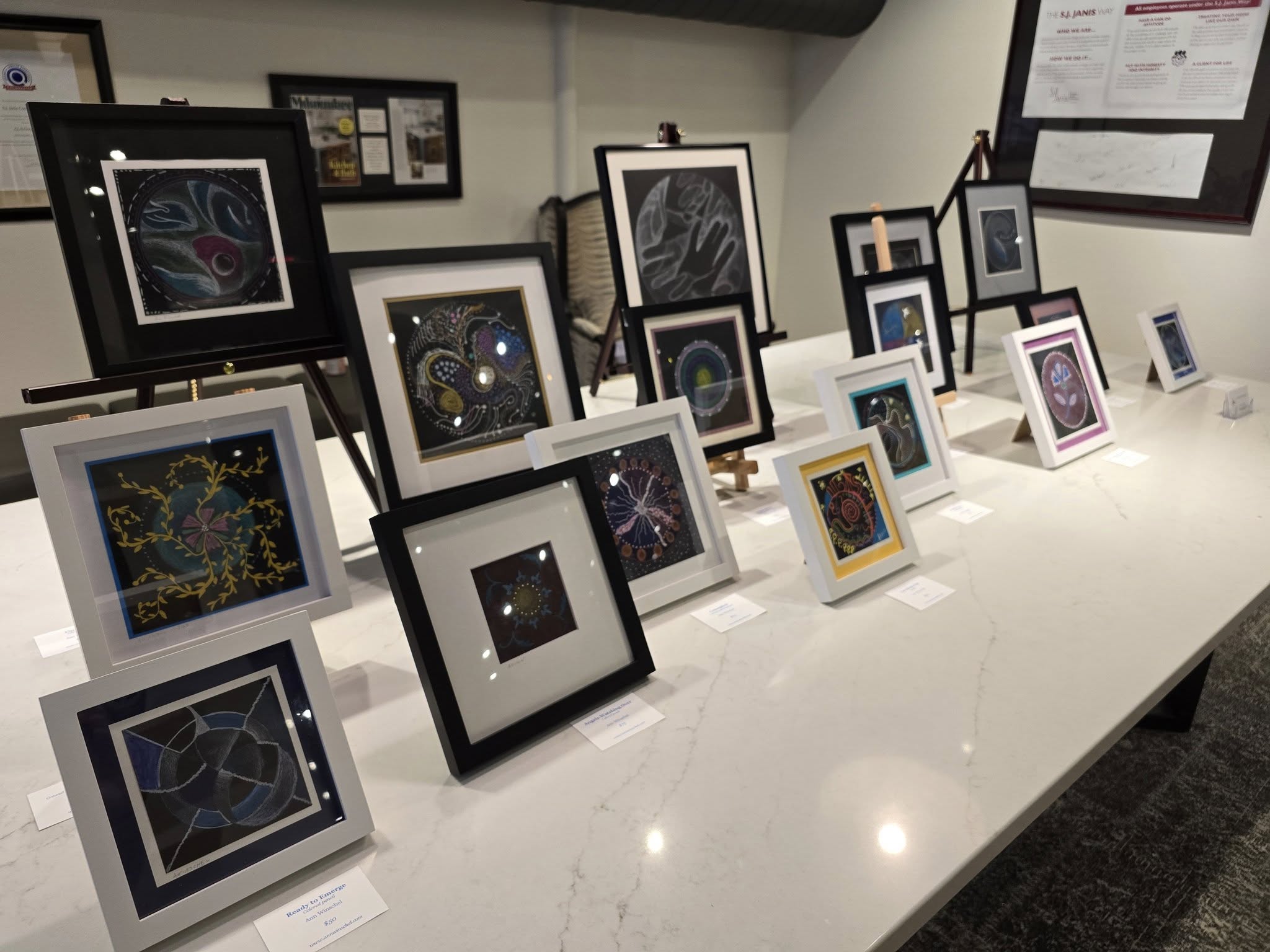 A variety of framed abstract artworks are displayed on a white table in a well-lit room, with informational pamphlets on the wall.