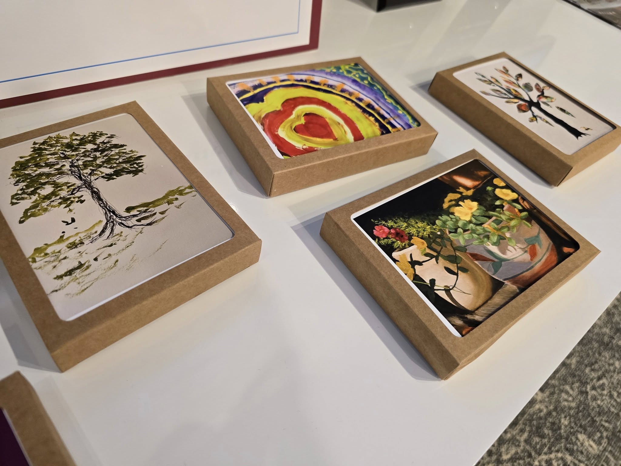 Four colorful paintings in cardboard frames, featuring nature themes like trees and flowers, are displayed on a white surface.