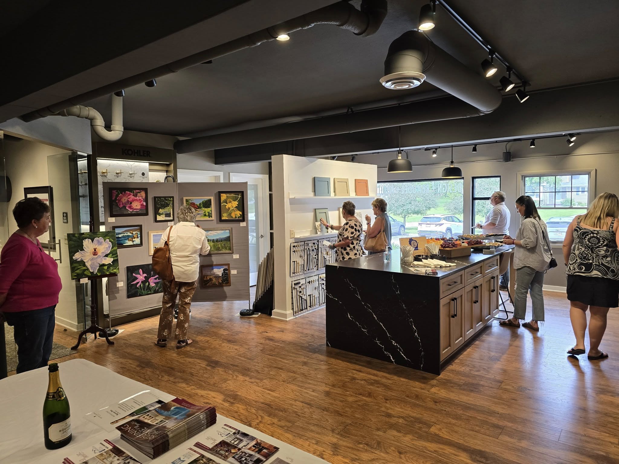 An art gallery with people observing paintings; art supplies and refreshments on display around a sleek, modern interior with wooden flooring.
