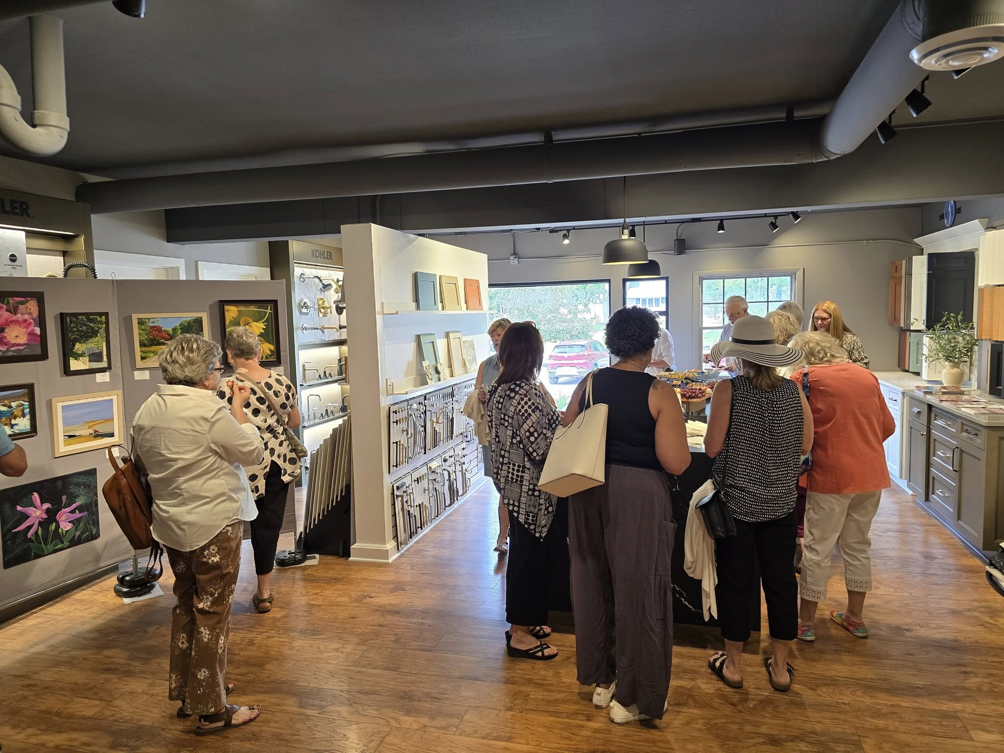 A group of people gather in an art exhibit, observing paintings displayed on the walls of a warmly lit room.