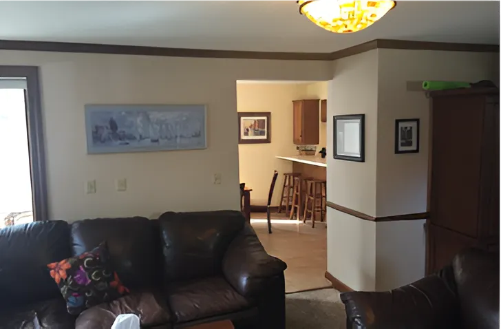 Cozy living room with brown leather sofas, a colorful pillow, and view into kitchen with bar stools. Warm lighting and framed art.