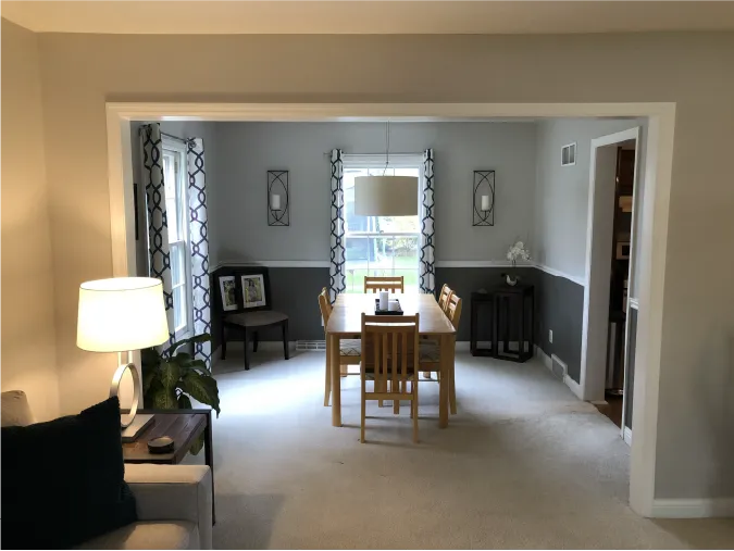 A cozy dining room with a wooden table and chairs, pendant light, decorative curtains, and neutral walls. Adjacent living space with a lamp.