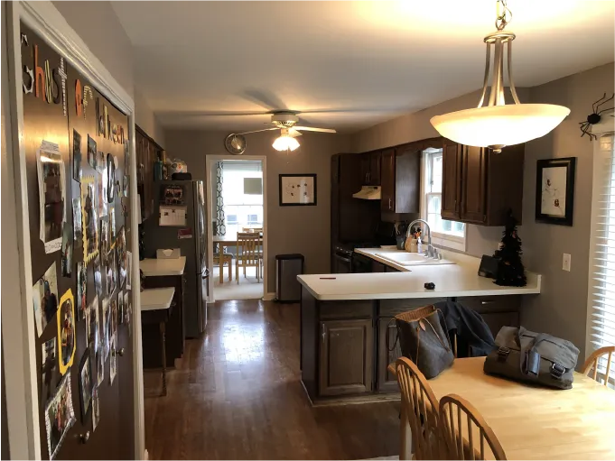 A kitchen with brown cabinets, white countertops, dining area, and wall decorations. Ceiling fan and lights create a cozy atmosphere.