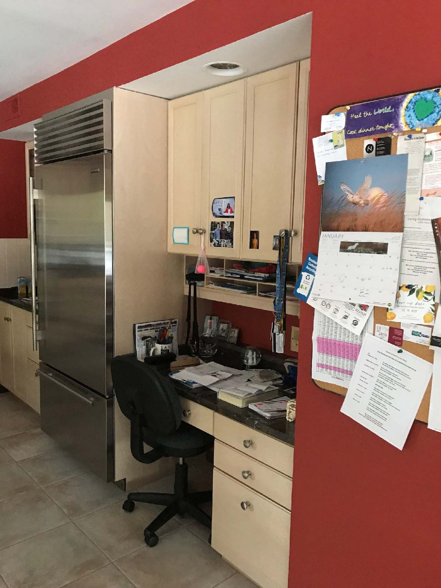A small home office nook with a desk, chair, calendars, and notes pinned on a vibrant red wall near a kitchen area.