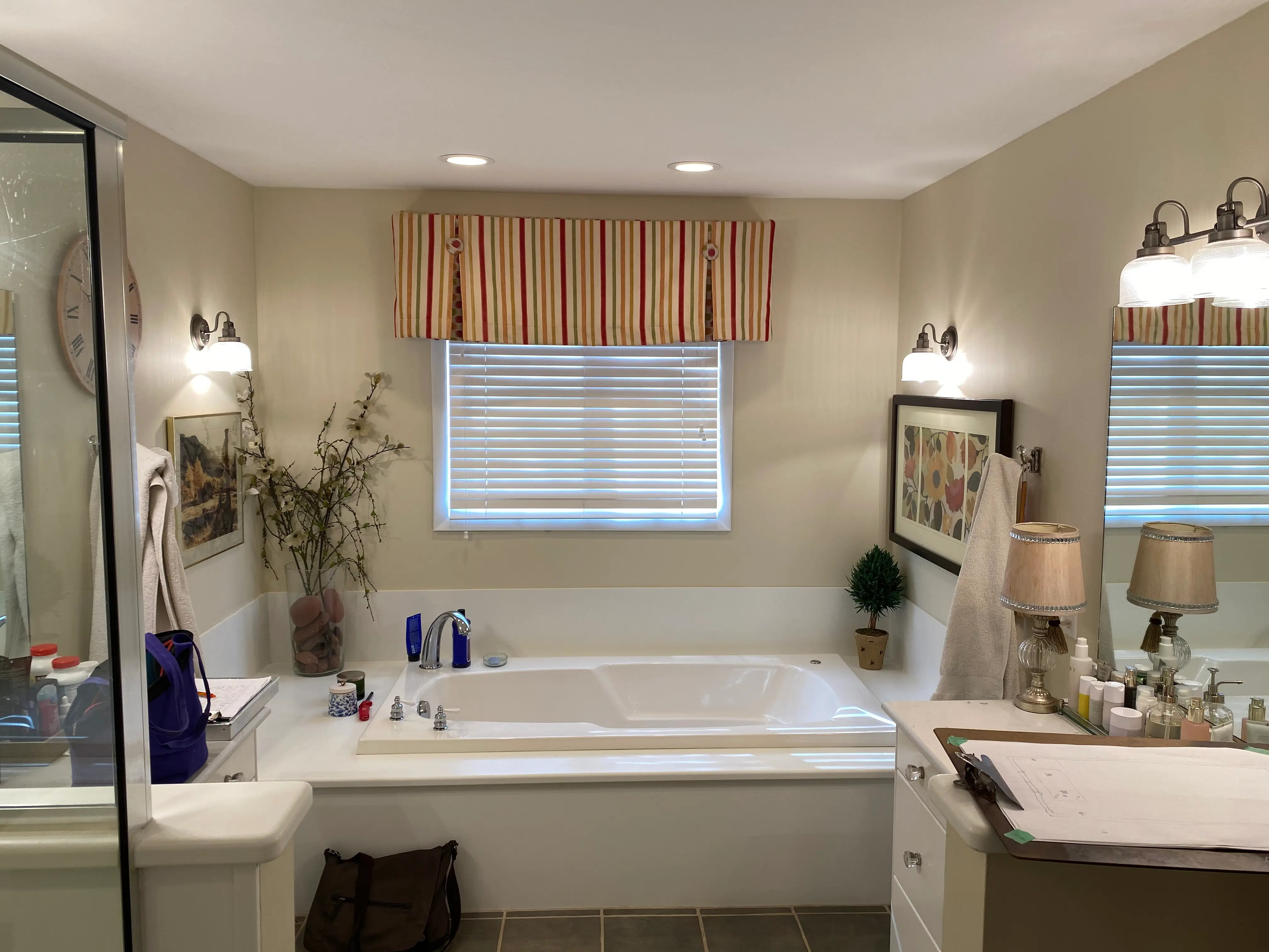 A cozy bathroom featuring a large bathtub, striped window valance, wall art, and various toiletries on the countertop under warm lighting.