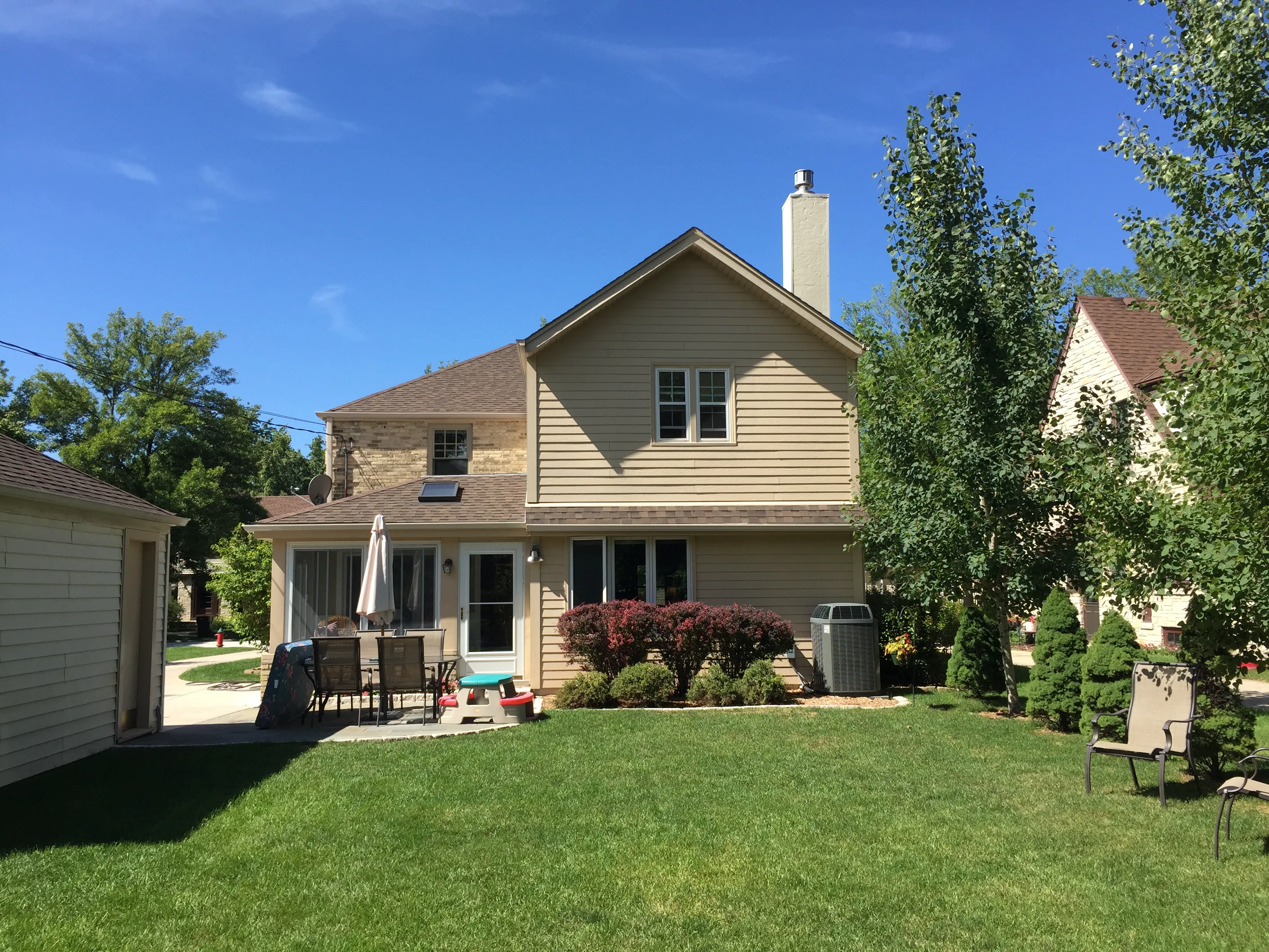 A suburban house with a spacious backyard, patio table, and chairs under clear blue skies. Trees and shrubs surround the well-manicured lawn.
